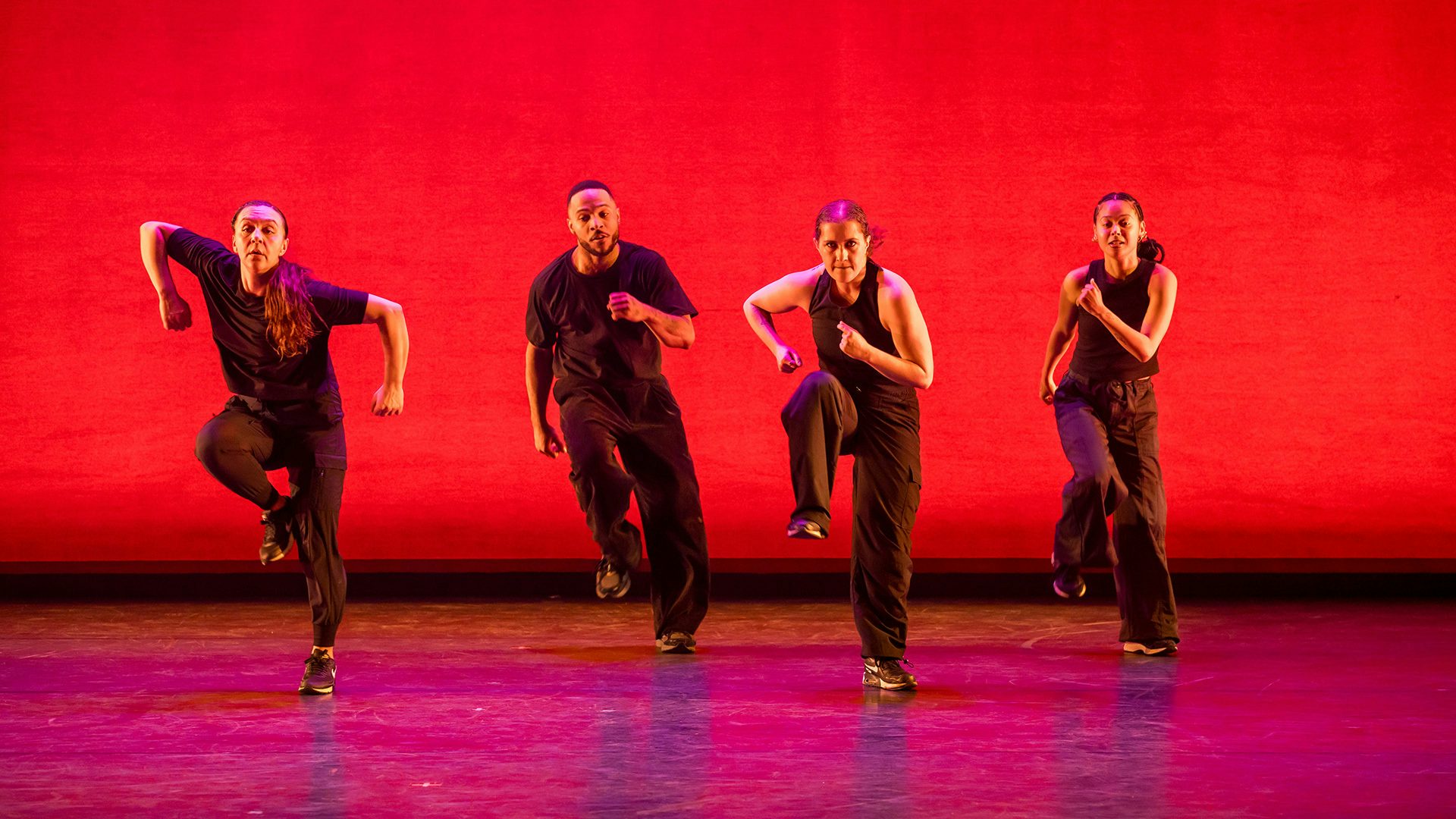 Four dancers wearing black clothes stand side by side on their left legs against a red background.