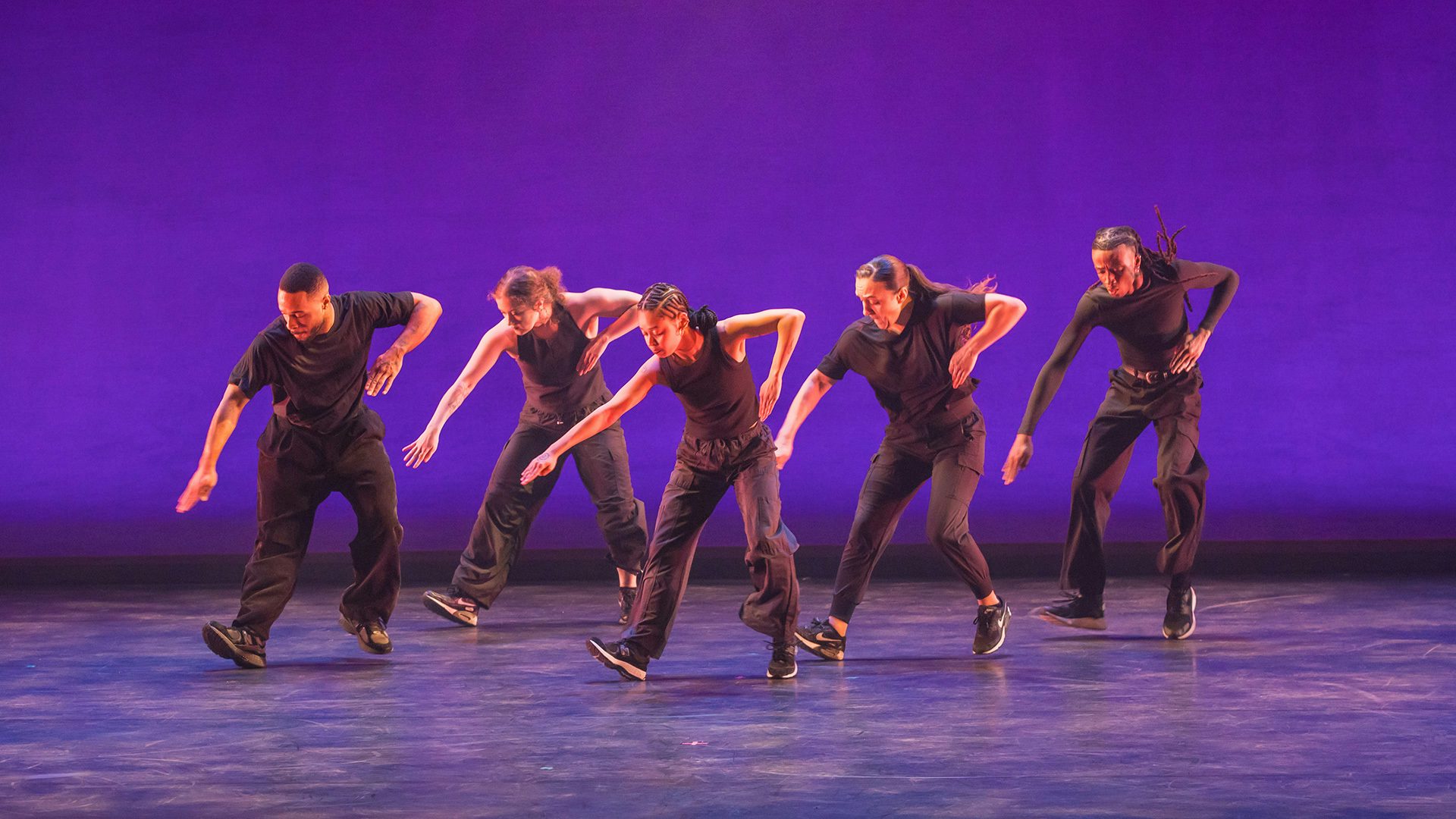 Five dancers wearing black clothes stand in a staggered line with their right arms pointed to the ground and their left arms bent. They dance against an indigo background.