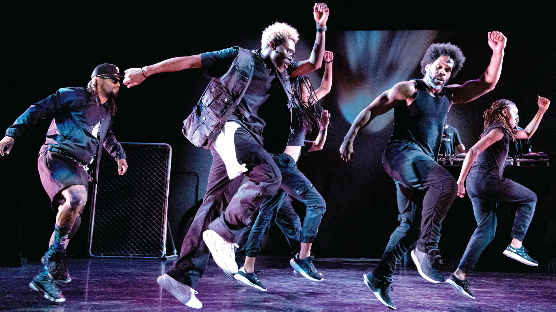 Five black dancers wearing black clothes are in the middle of an energetic step with their arms in the air while someone controls a sound system behind them.