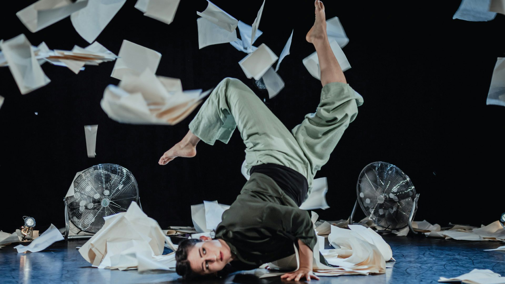 A dancer is twisted with her head on the ground and her legs in the air while two fans blow blank papers around her.