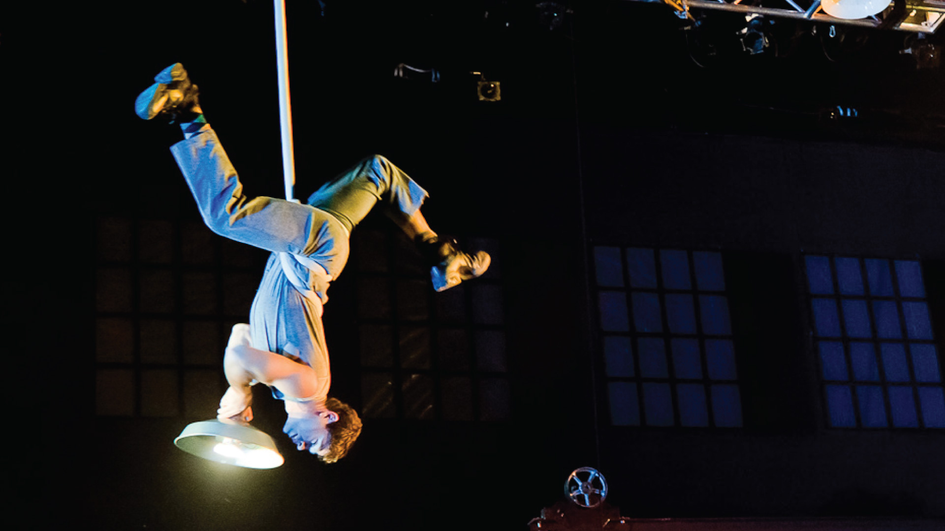 A man wearing a gray jumpsuit hangs upside down with his legs outstretched in the air and has a hanging light wrapped around his waist.