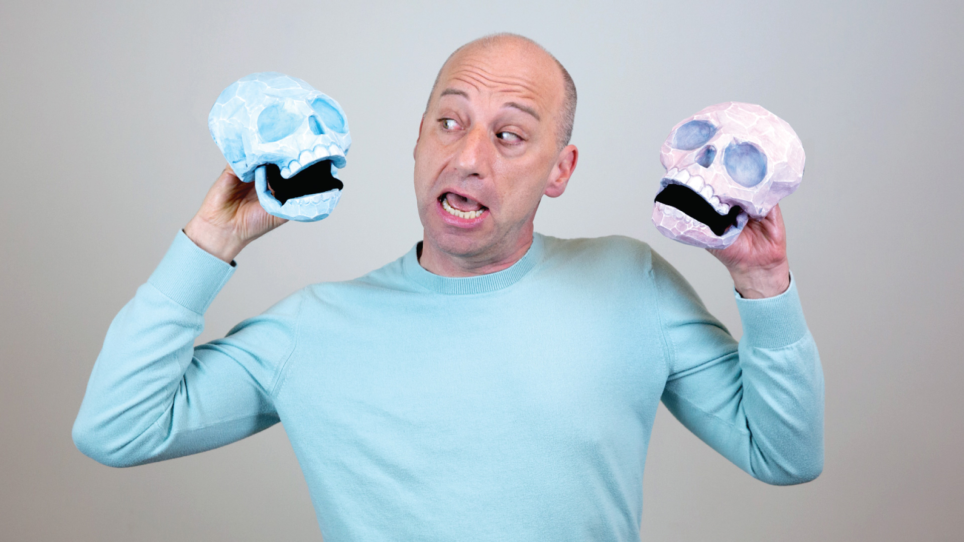 A man in a light blue, long sleeve sweater holds one skull in each hand, one that's blue and one that's pink, against a gray background.