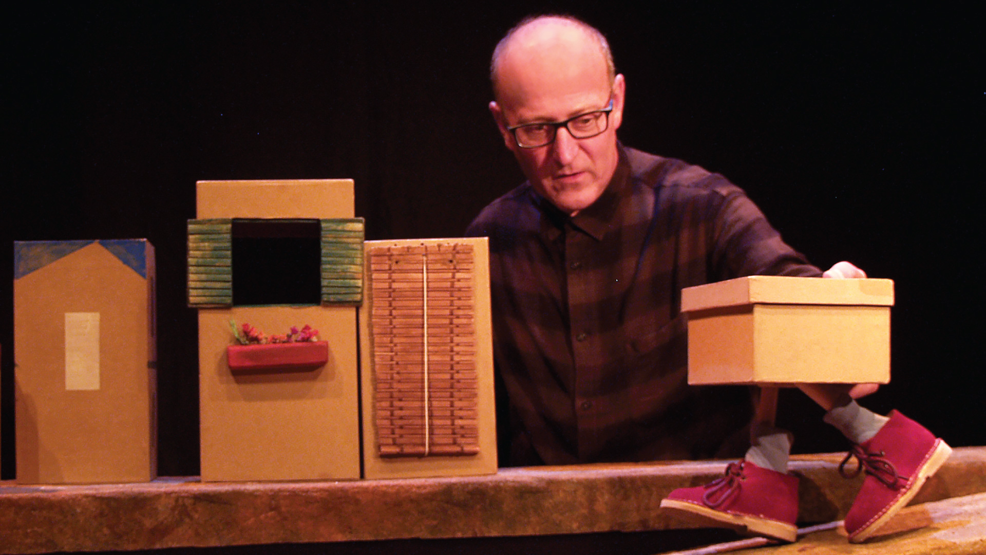 A man with glasses sitting next to three small, cardboard buildings puppeteers a small shoe box that has a pair of short legs, white socks and red shoes.