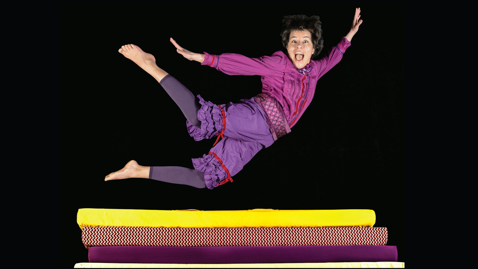 A performer with short, dark hair and purple clothes is stretched out in the middle of a jump hovering above several thin mattresses.