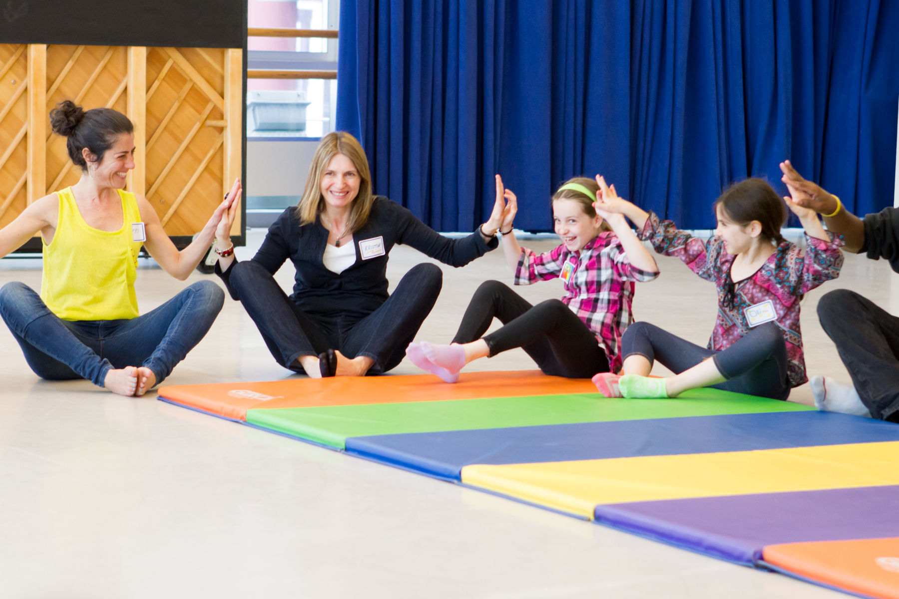 Two adults and two kids sit on a colorful mat on the ground while touching their hands together.