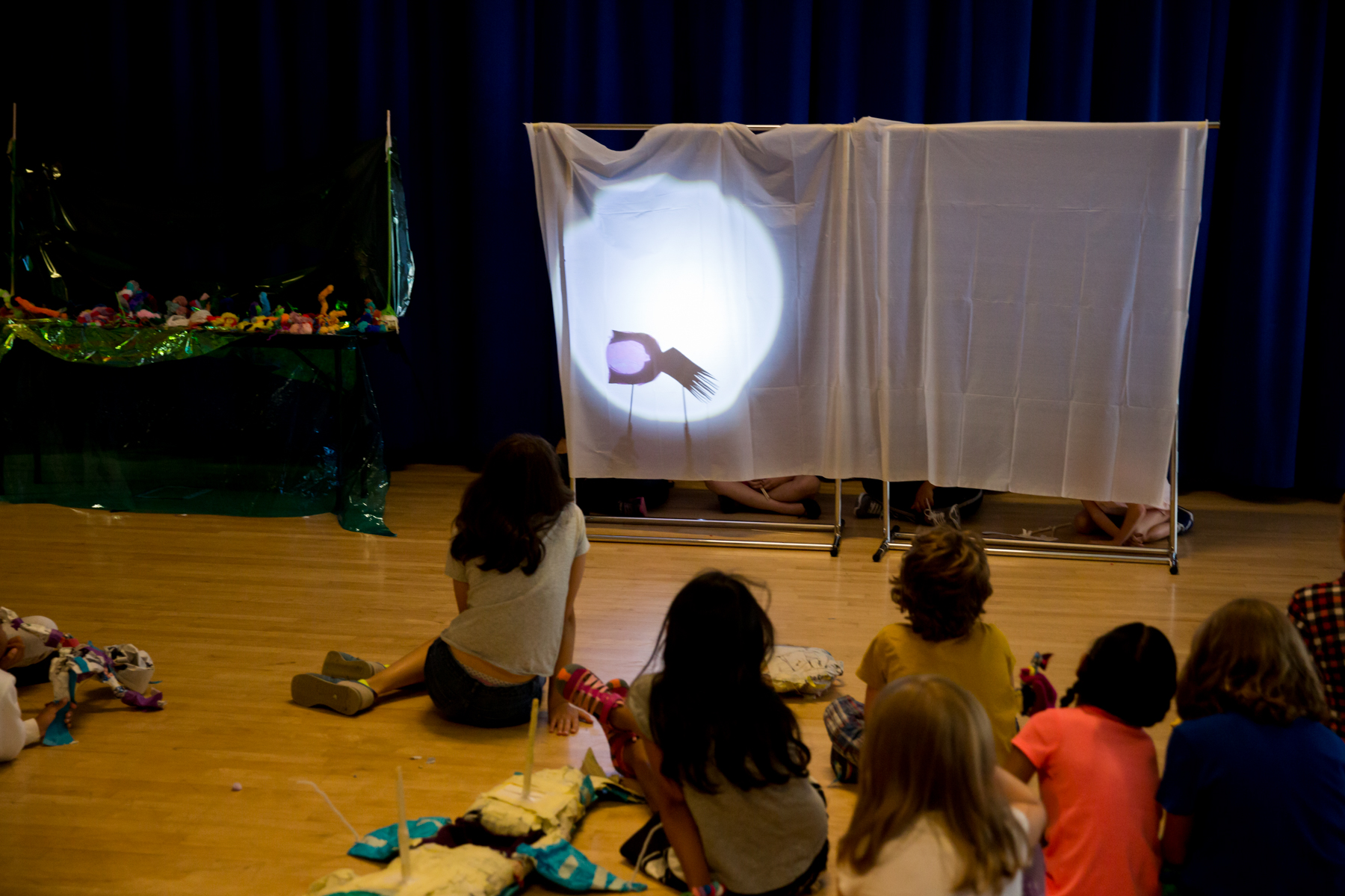 A group of people on the ground watch a shadow puppet performance on a white screen, with a table full of puppetry materials to the left of the screen.