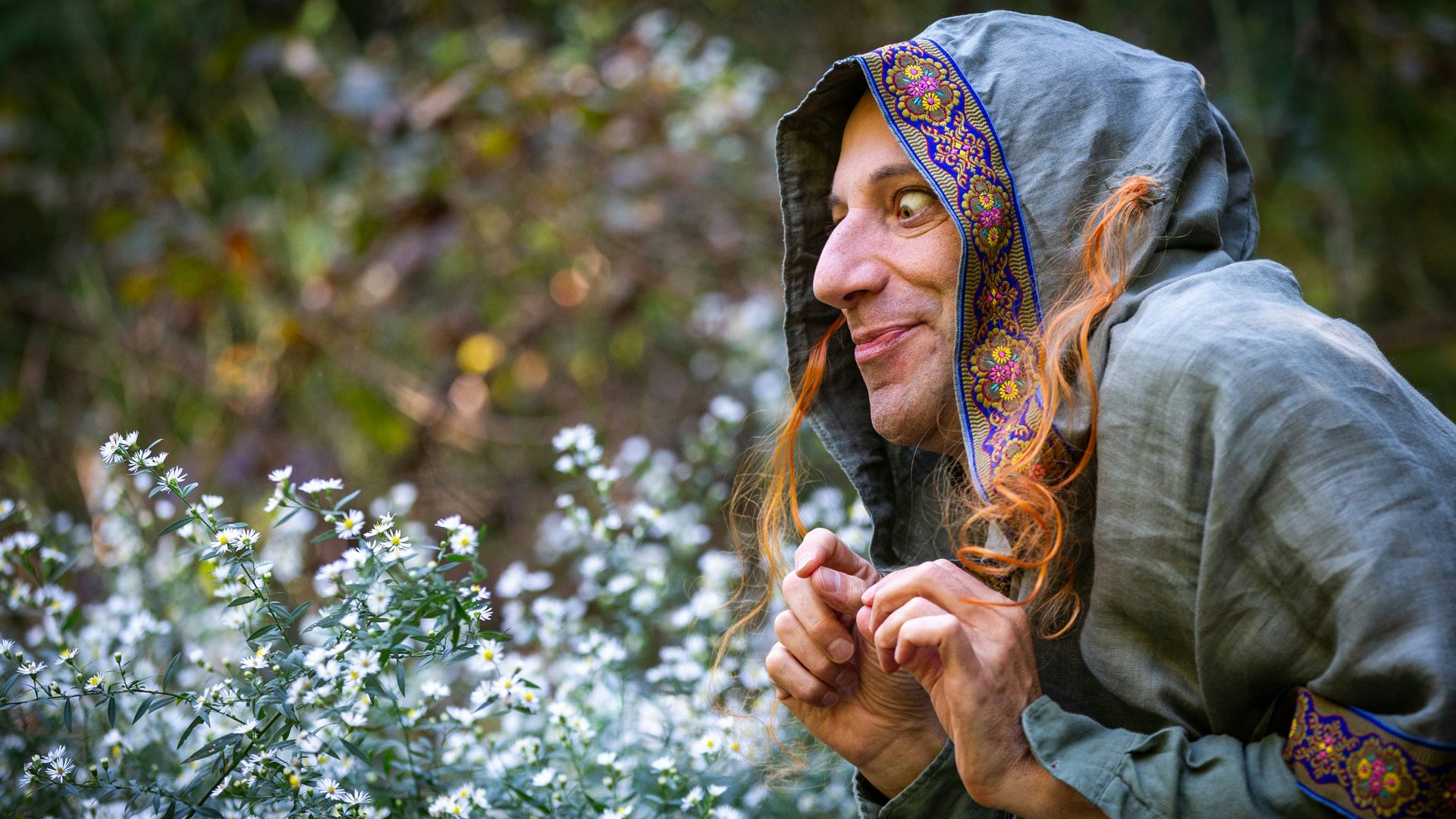 A person wearing a gray hood with orange hair coming out of it crouches and looks to the left in a field of flowers.