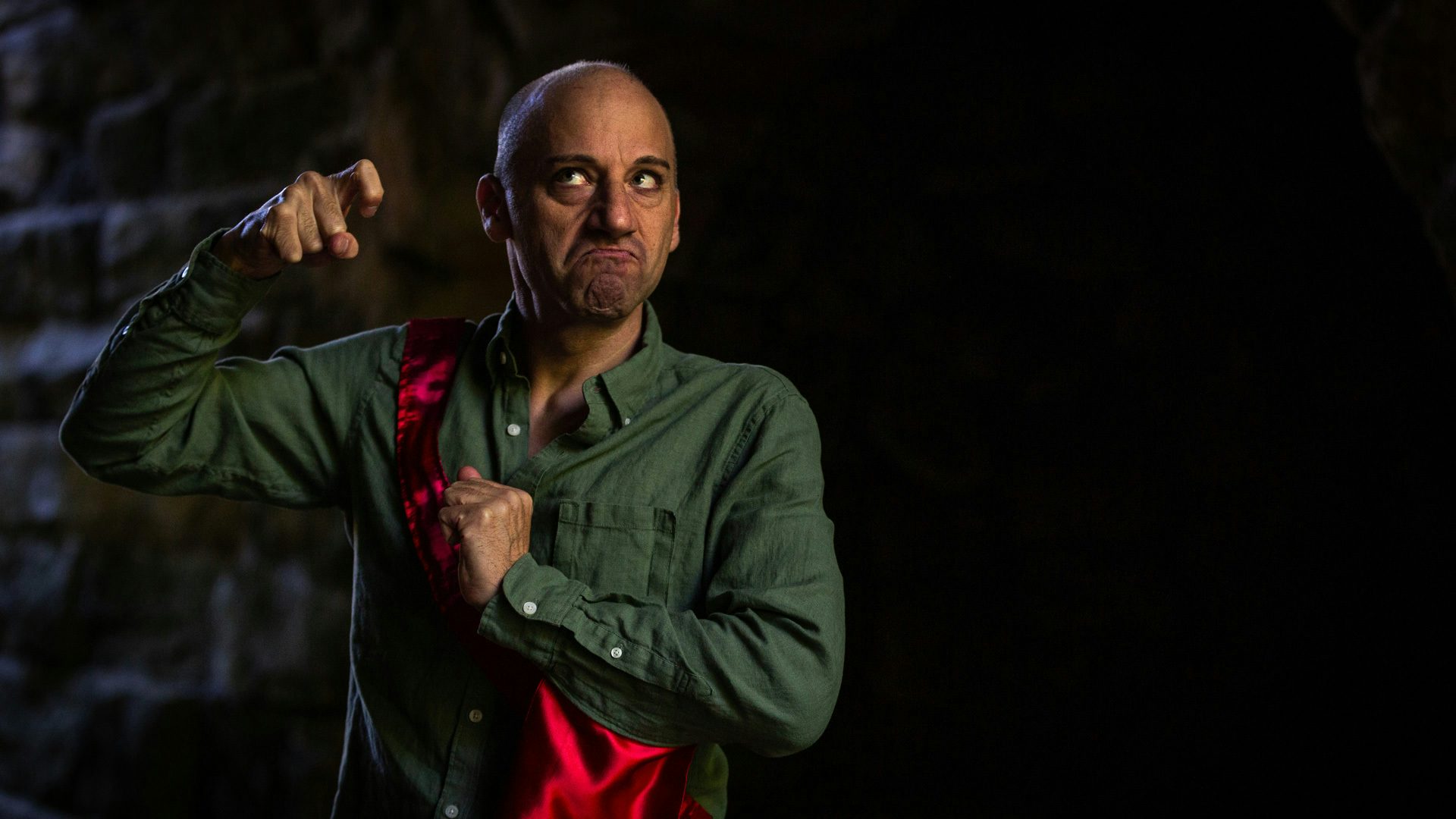 A man wearing a green shirt frowns and looks up and to the left at his hand which is in a hook shape.