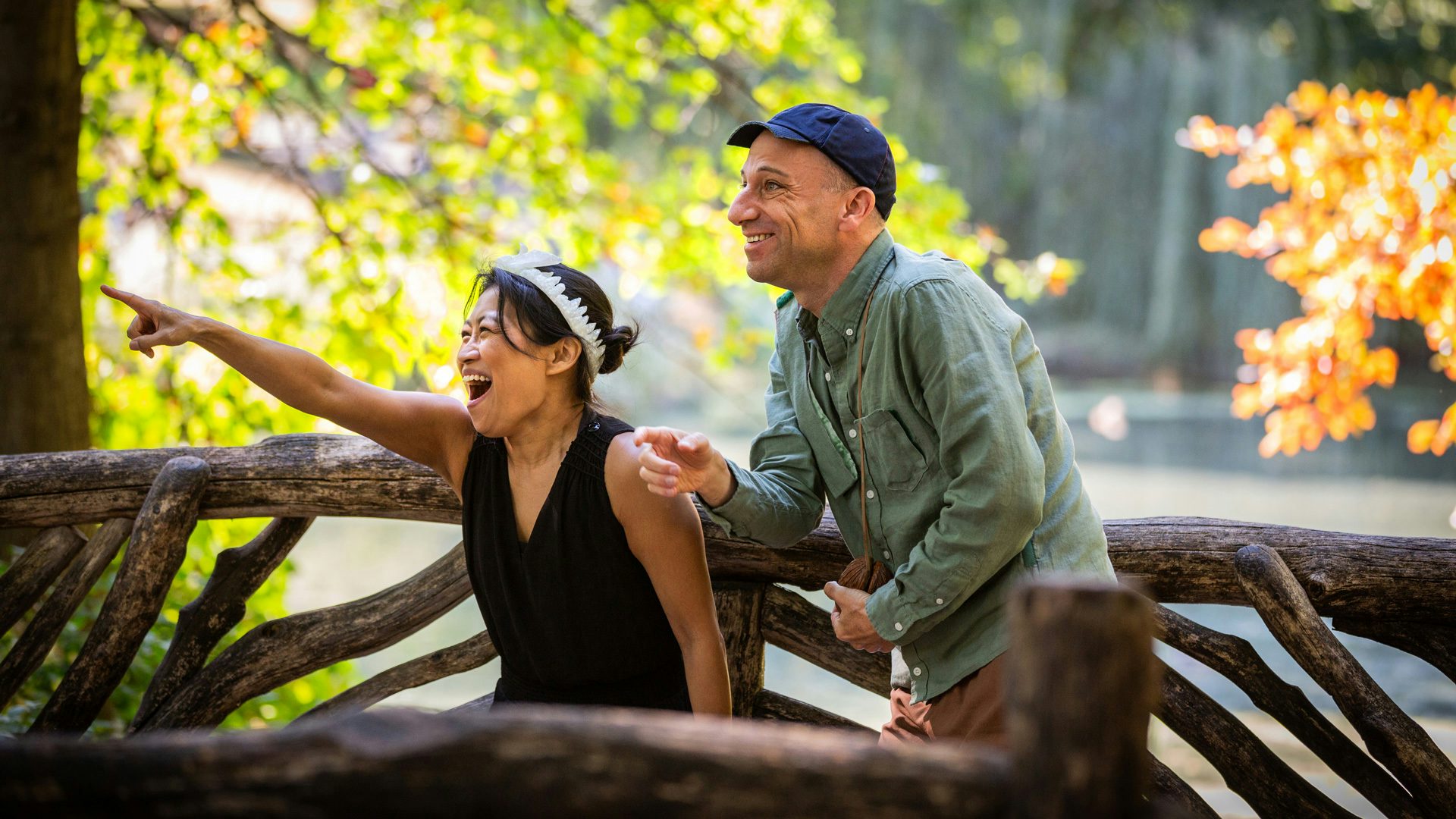 A woman wearing a black shirt and white headband points off to the left, and a man wearing a green shirt and navy hat next to her looks where she's pointing. They're on a wooden bridge in a forest.
