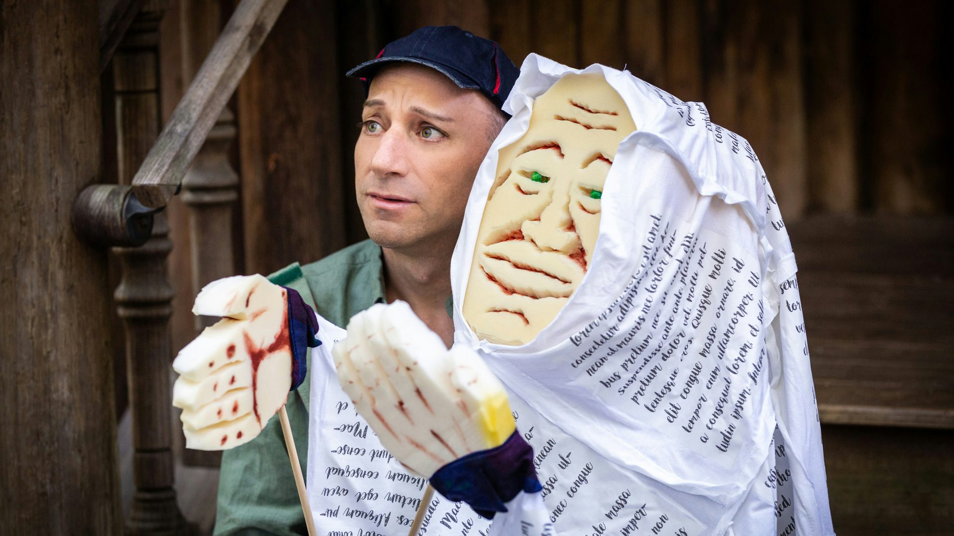 A man wearing a navy hat holds a puppet of an aged person covered in white clothes and hood with black writing on it.