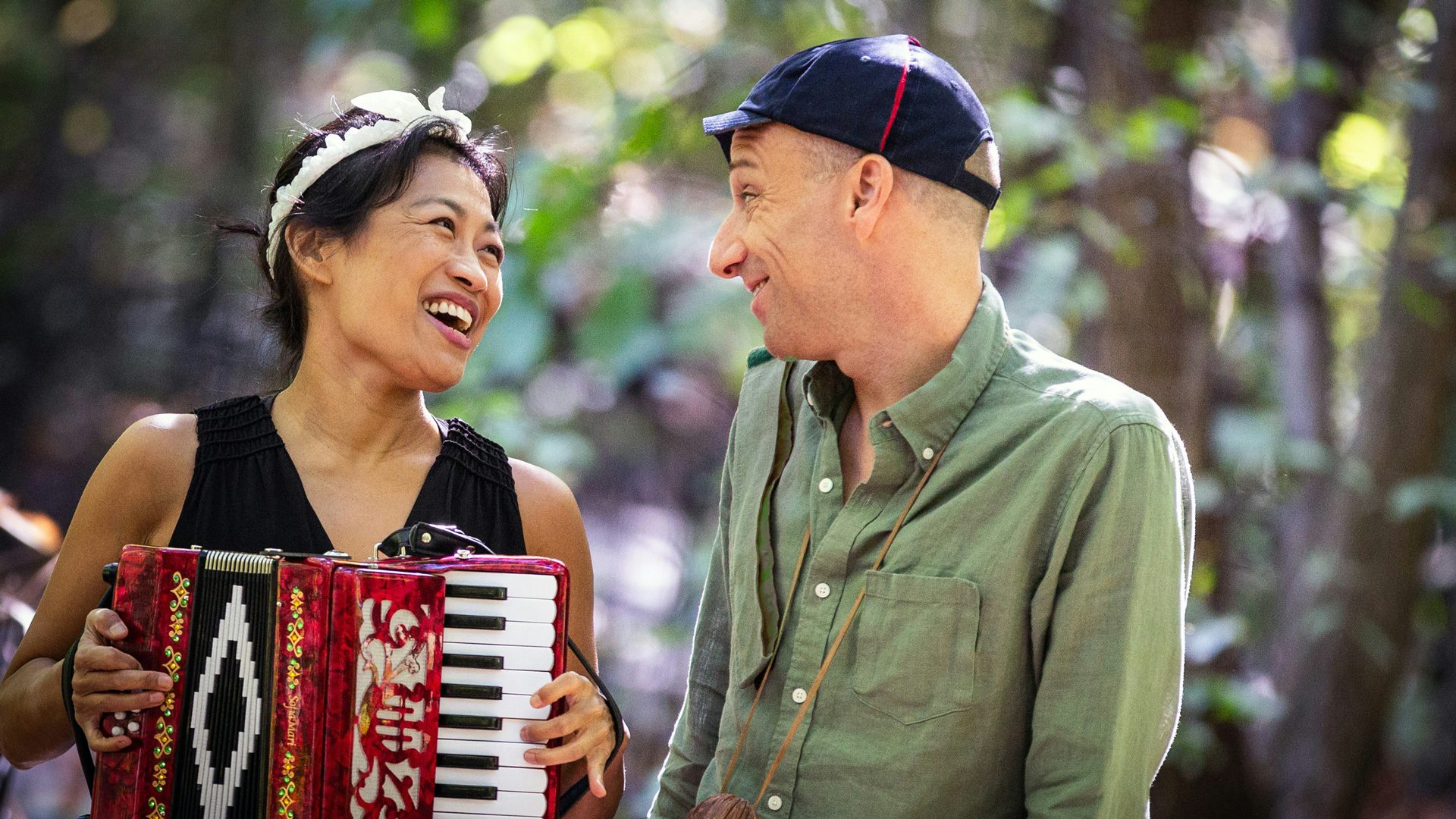 A woman wearing a black shirt and white headband plays an accordion and smiles at a man to the right of her, who wears a navy hat and green shirt.