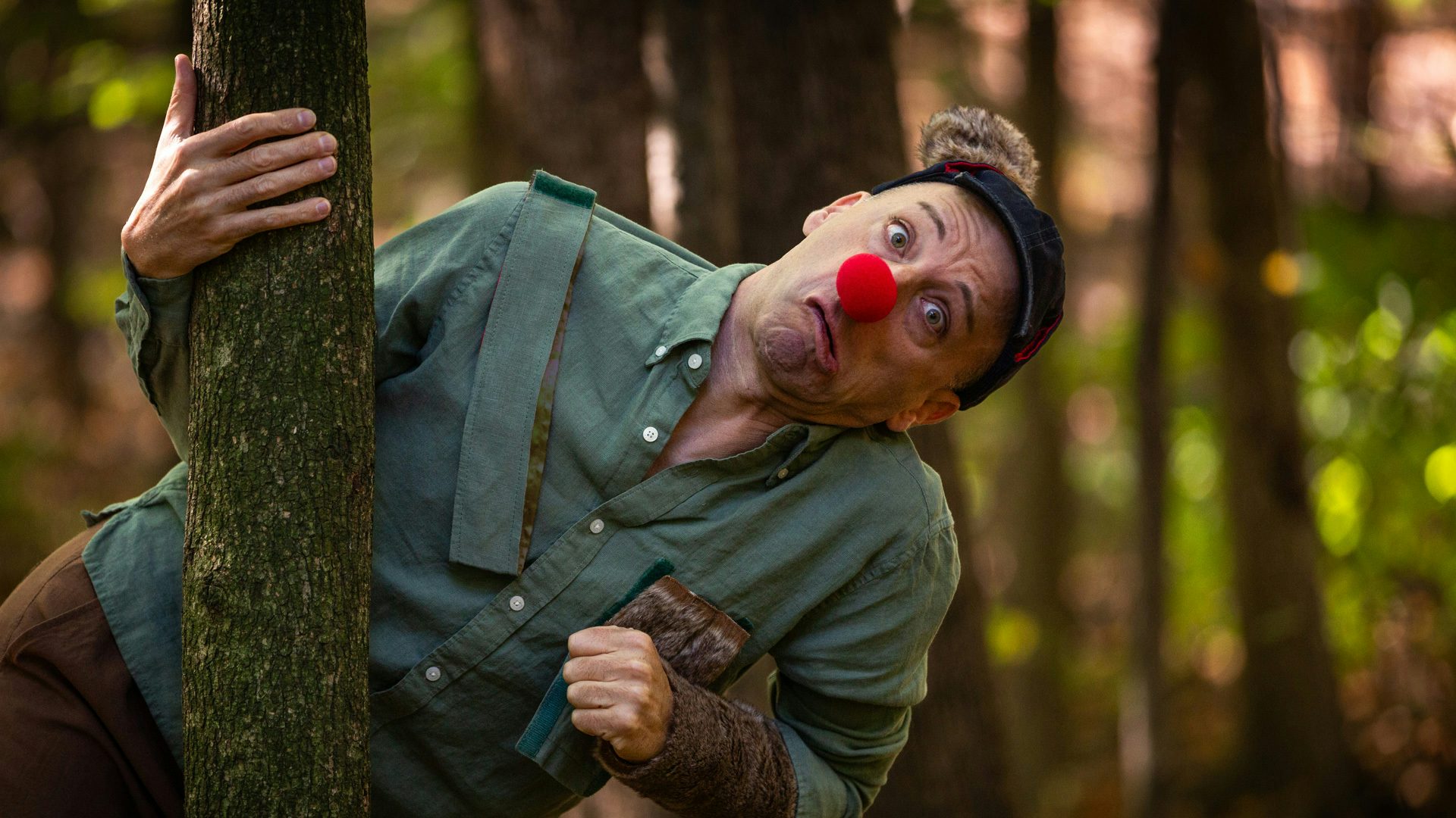 A man wearing a red clown nose frowns and leans to the right from behind a tree.