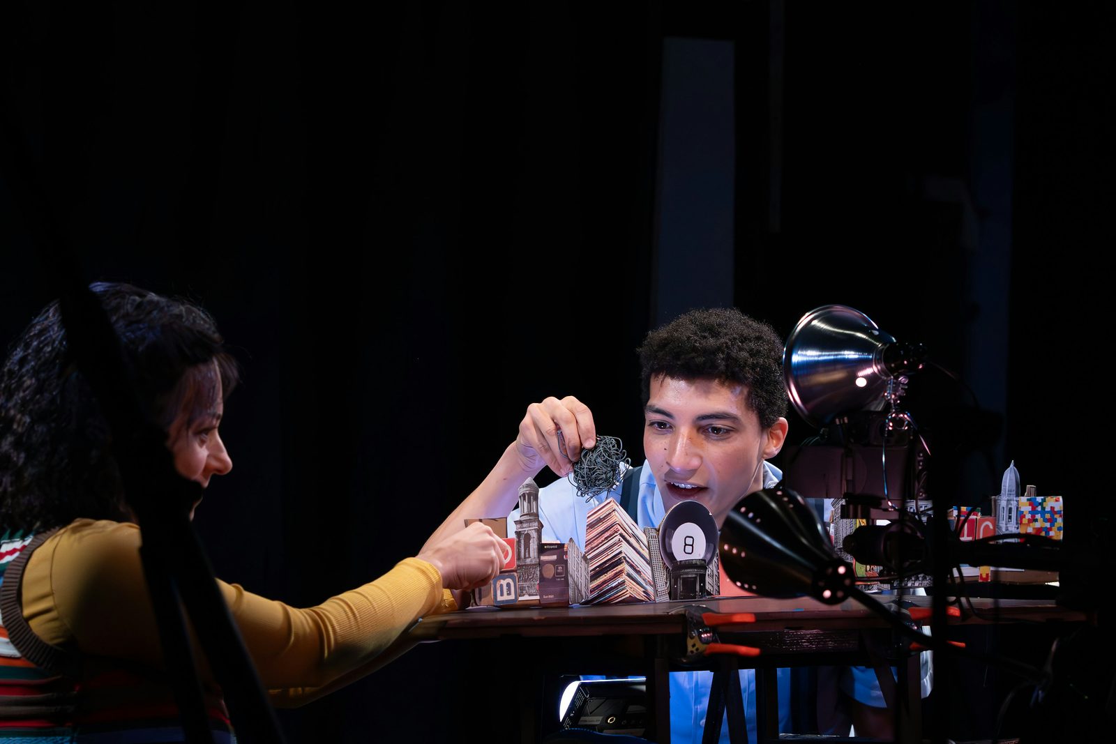 Two lamps shine on a boy who plays with miniatures of a city on a table. To the left, a girl watches and moves the miniatures around too.