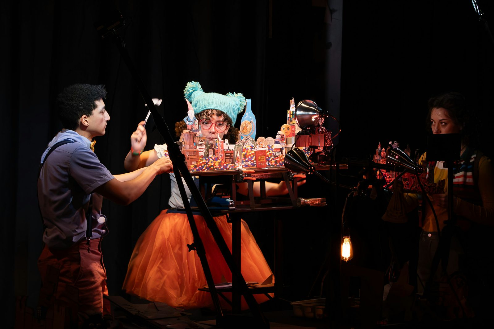 A girl wearing red glasses and a blue hat plays with a miniature city on a table with three lamps shining on it. On the left, a boy wearing a blue shirt plays along, and on the right, another person watches them.