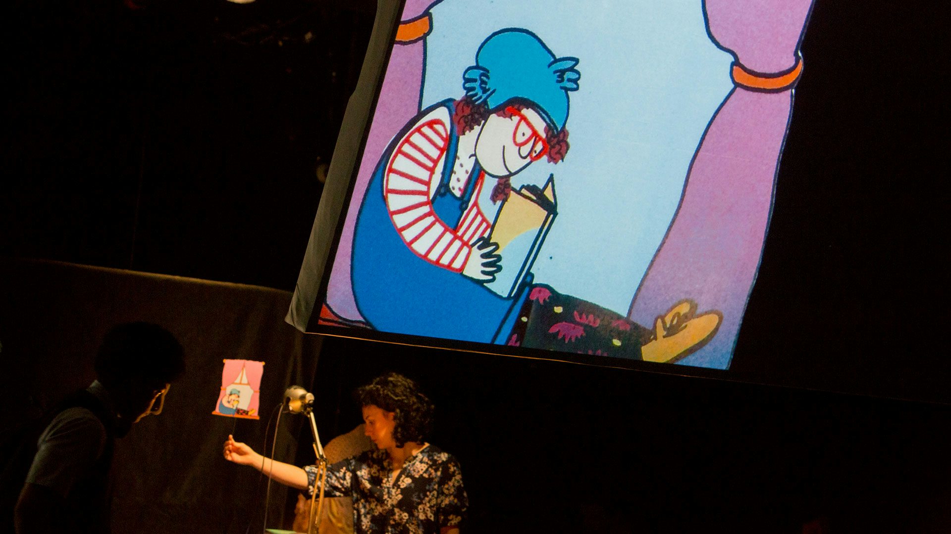 A woman holds up an image of a girl with bright red hair and glasses reading in a windowsill while the image is projected largely onto a screen above her.