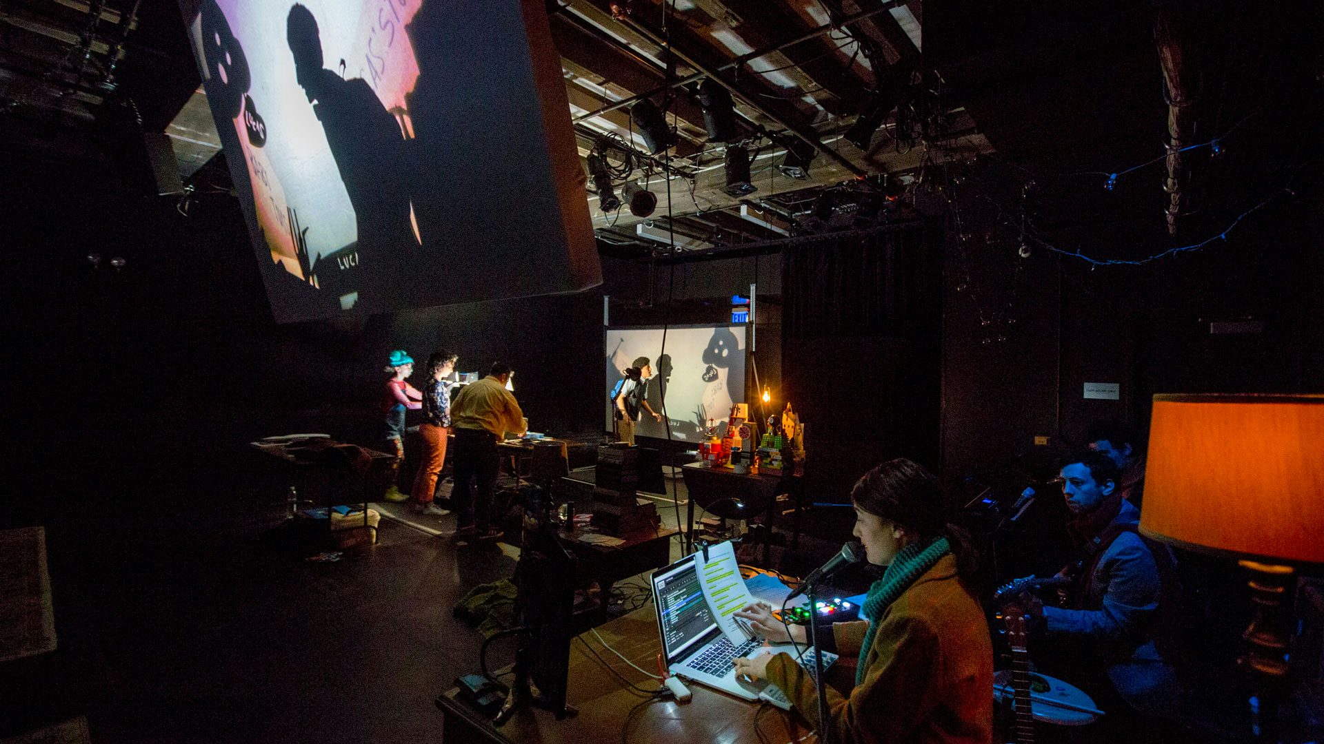 A behind-the-scenes look at the factors that go into the production: a stage manager reads cues into a microphone while three cast members create shadows for a projection screen and one actor interacts with the shadows on the screen.