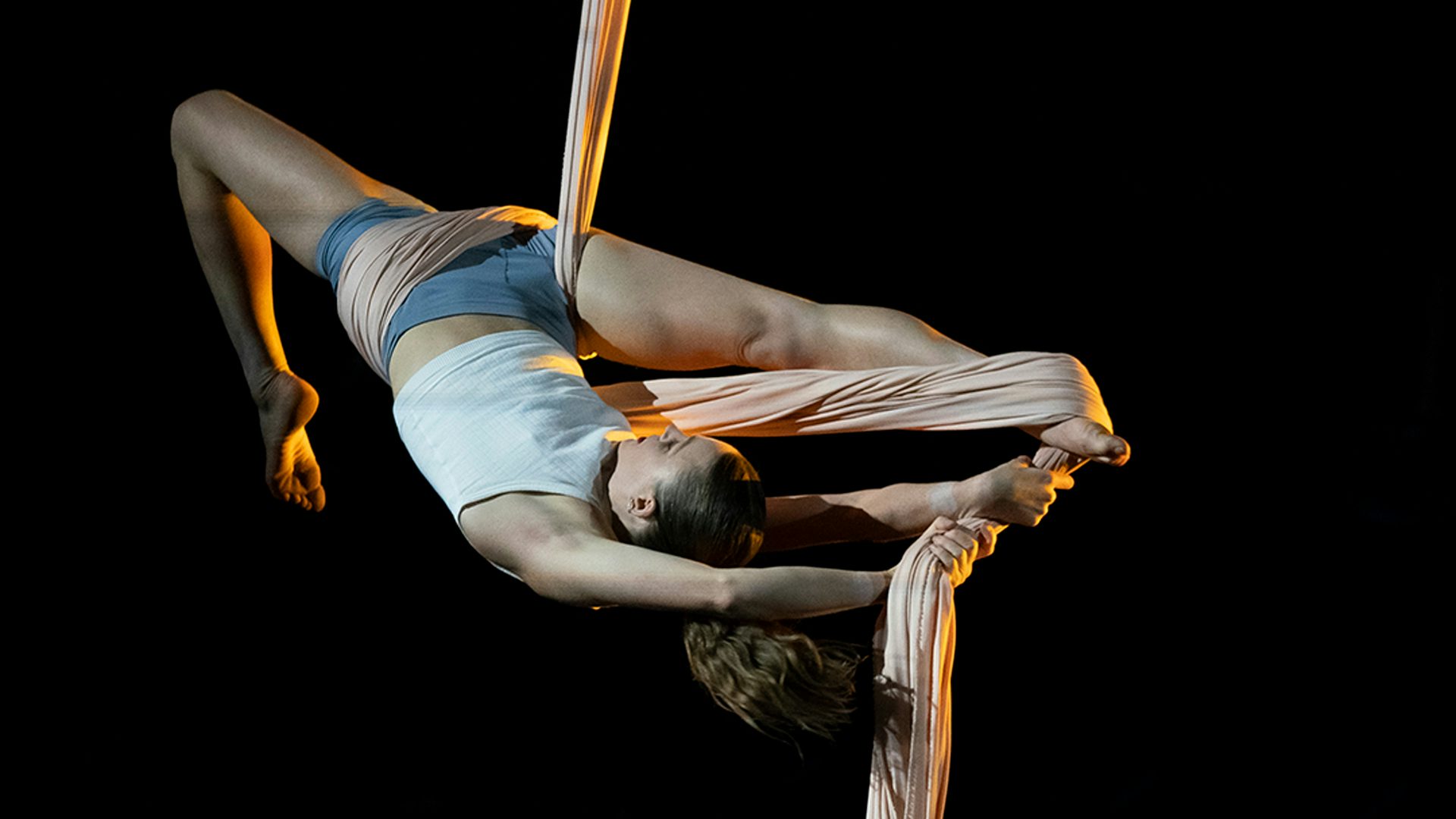 A girl hanging upside down in a pale pink piece of silk stretches her right leg toward her face as her left leg bends toward her back.