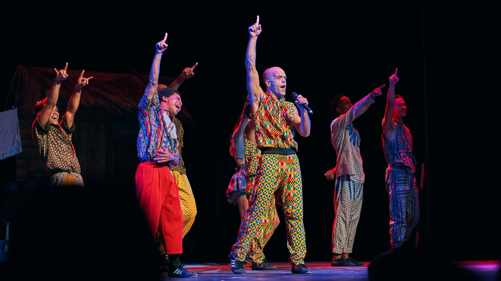 A group of performers wearing colorful clothing hold up their right hands and point toward the sky. A man in the middle performs with a microphone.