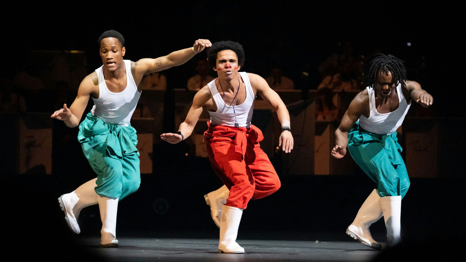 Three men wearing white tank tops, colorful pants and white rubber boots are in the middle of doing a gumboot dance step.