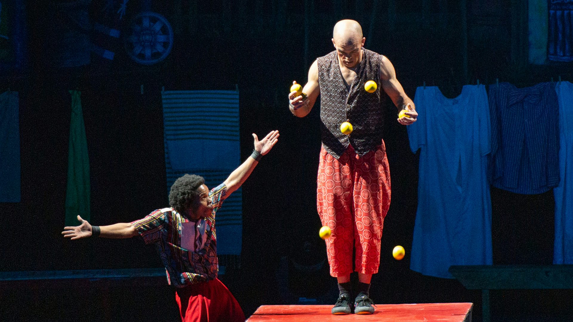 A bald man juggles six yellow balls while standing on a red platform, and another man with dark, curly hair gestures to him below the platform.