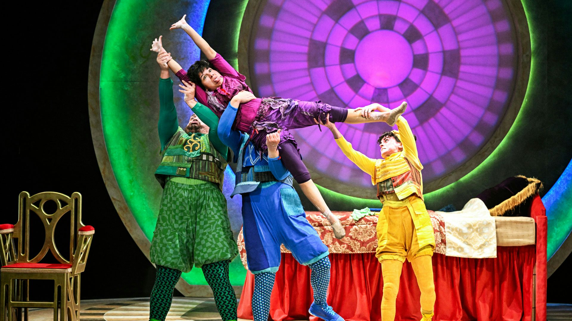 Three performers wearing green, blue and yellow clothes hold up a fourth performer wearing purple clothes who stretches towards the left side of the photo in front of a prop mattress, chair and circular backdrop.