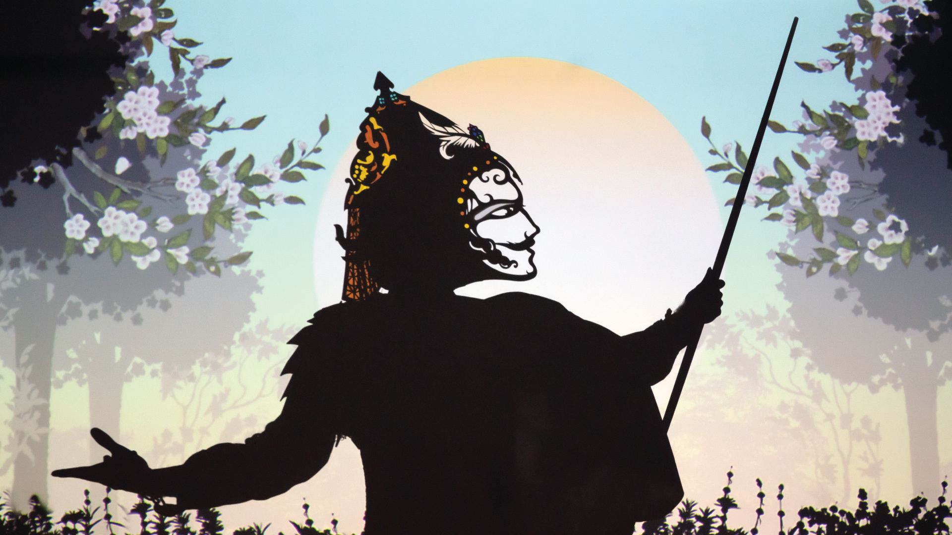 A performer wears a headpiece that casts a shadow of a face against a projection of trees and the sun.
