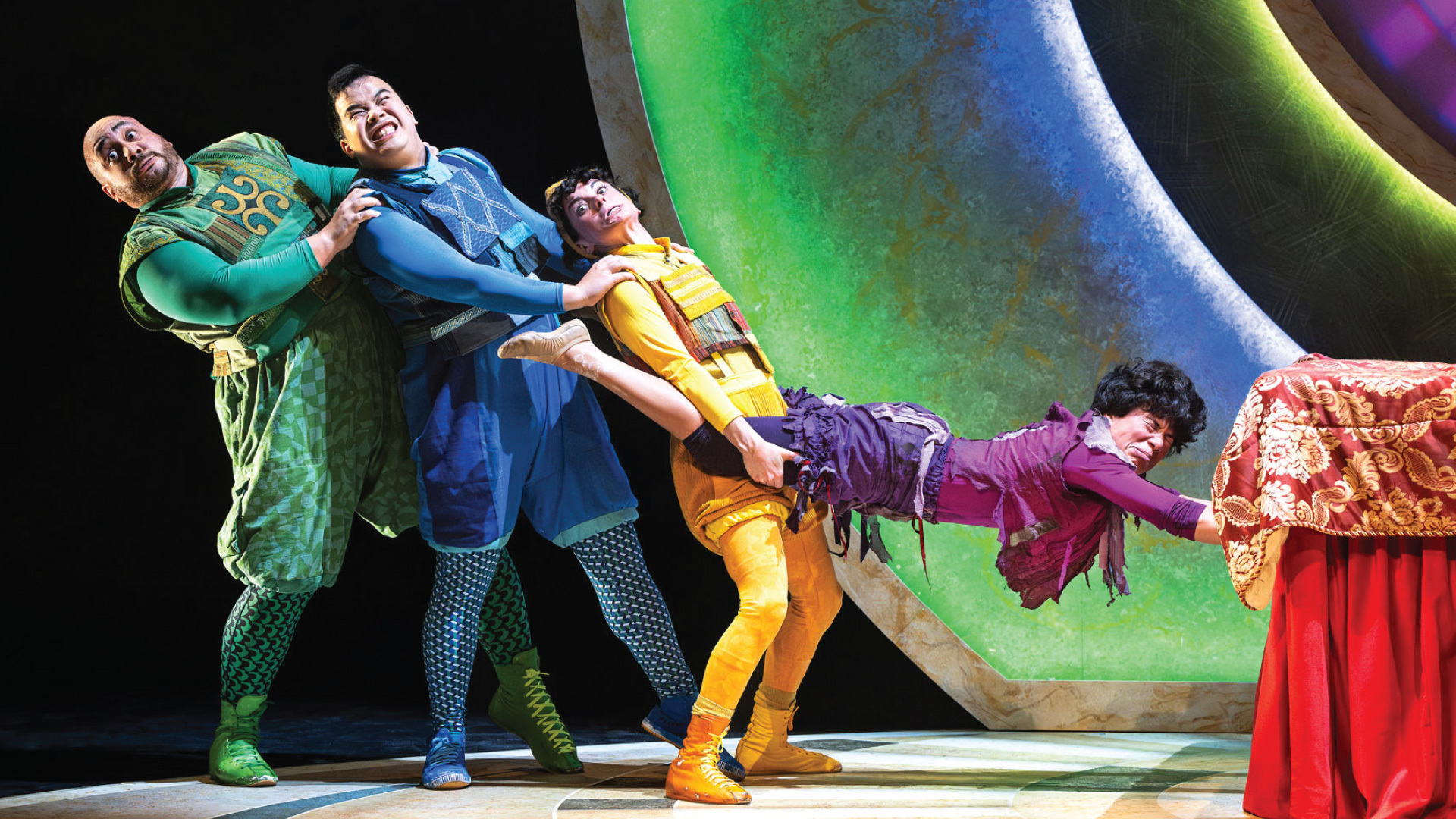 Three performers wearing green, blue and yellow clothes stand one in front of the other and work hard to pull a fourth performer wearing purple clothes away from the end of a bed.