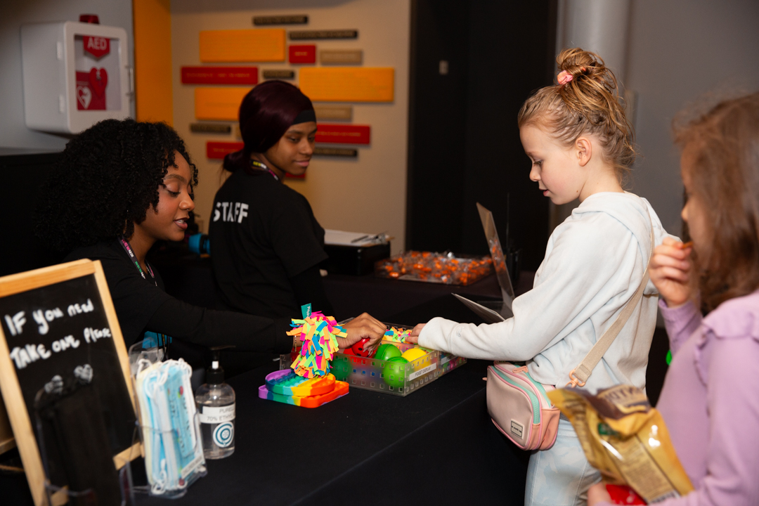 Two ushers at the Accessibility Table giving fidget toys to young patrons.