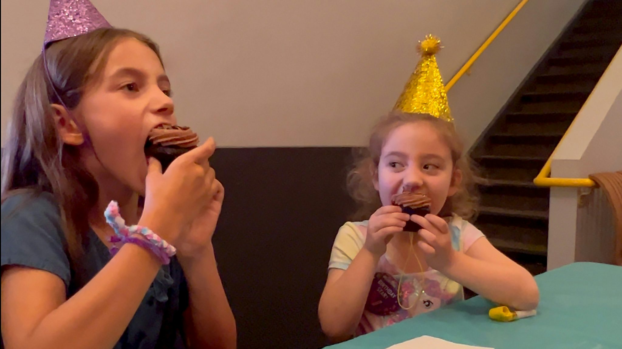 Two girls wearing party hats eat chocolate cupcakes.