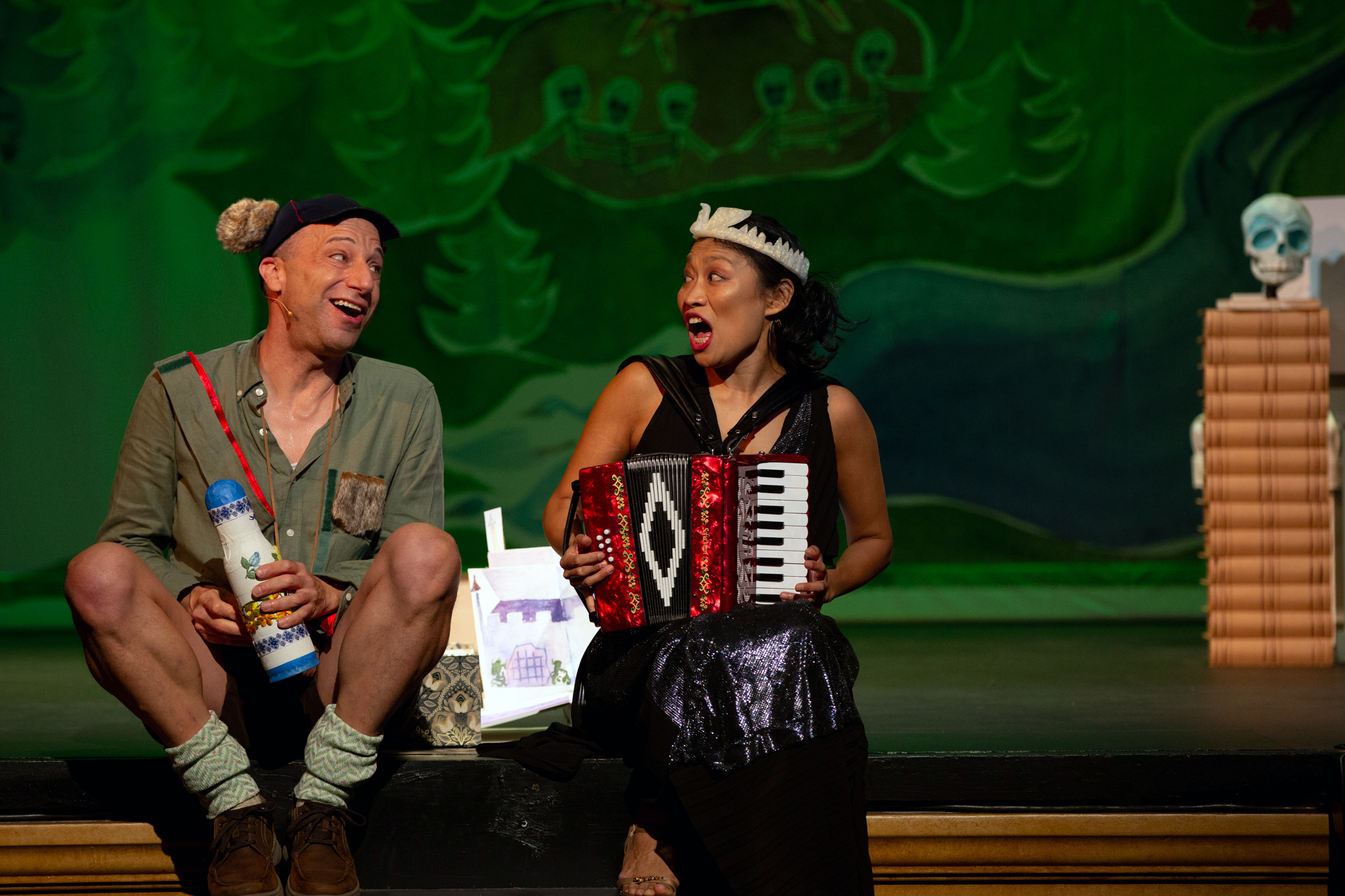 Anton Dudley and Faye Chiao sit on the edge of the stage singing while Faye plays accordion