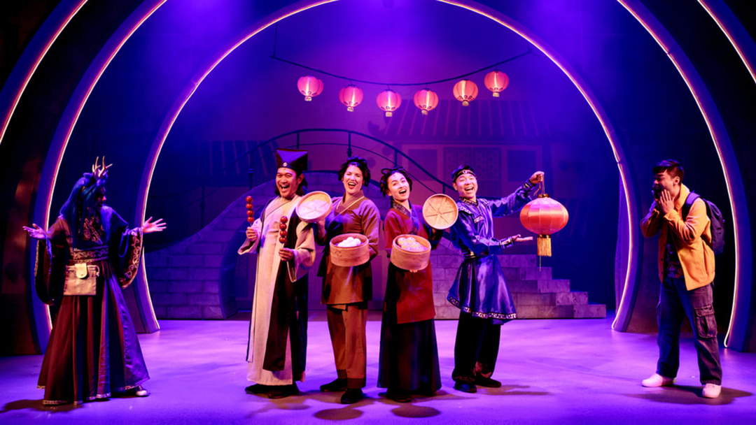 Five performers in colorful traditional costumes hold food items and lanterns on a stage lit with vibrant purple light.