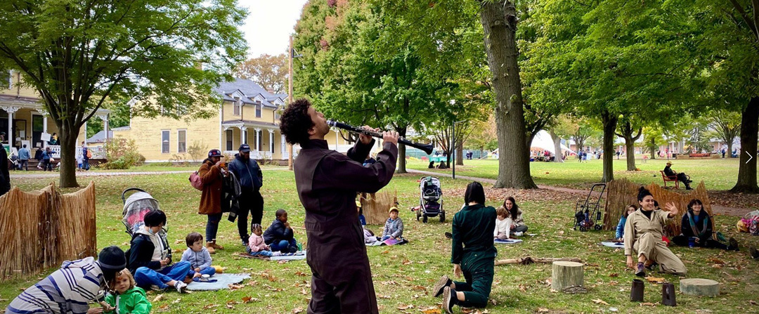 A performer plays a clarinet in a park surrounded by scattered audience members sitting on blankets and tree stumps.