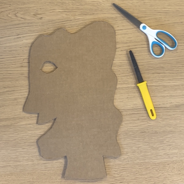 A cutout cardboard profile of a smiling face with wavy hair, alongside scissors and a cardboard cutter 