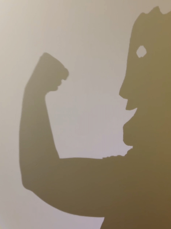 A shadow on a white wall of the mask atop a human body flexing their arm