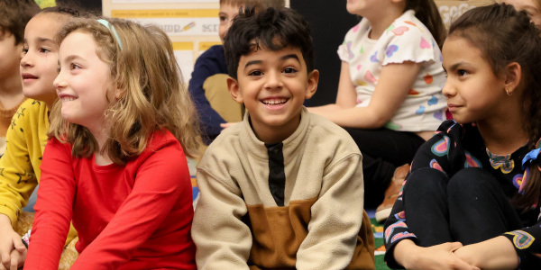 Kids smiling and one kid smiling while looking at the camera wearing a brown sweater.