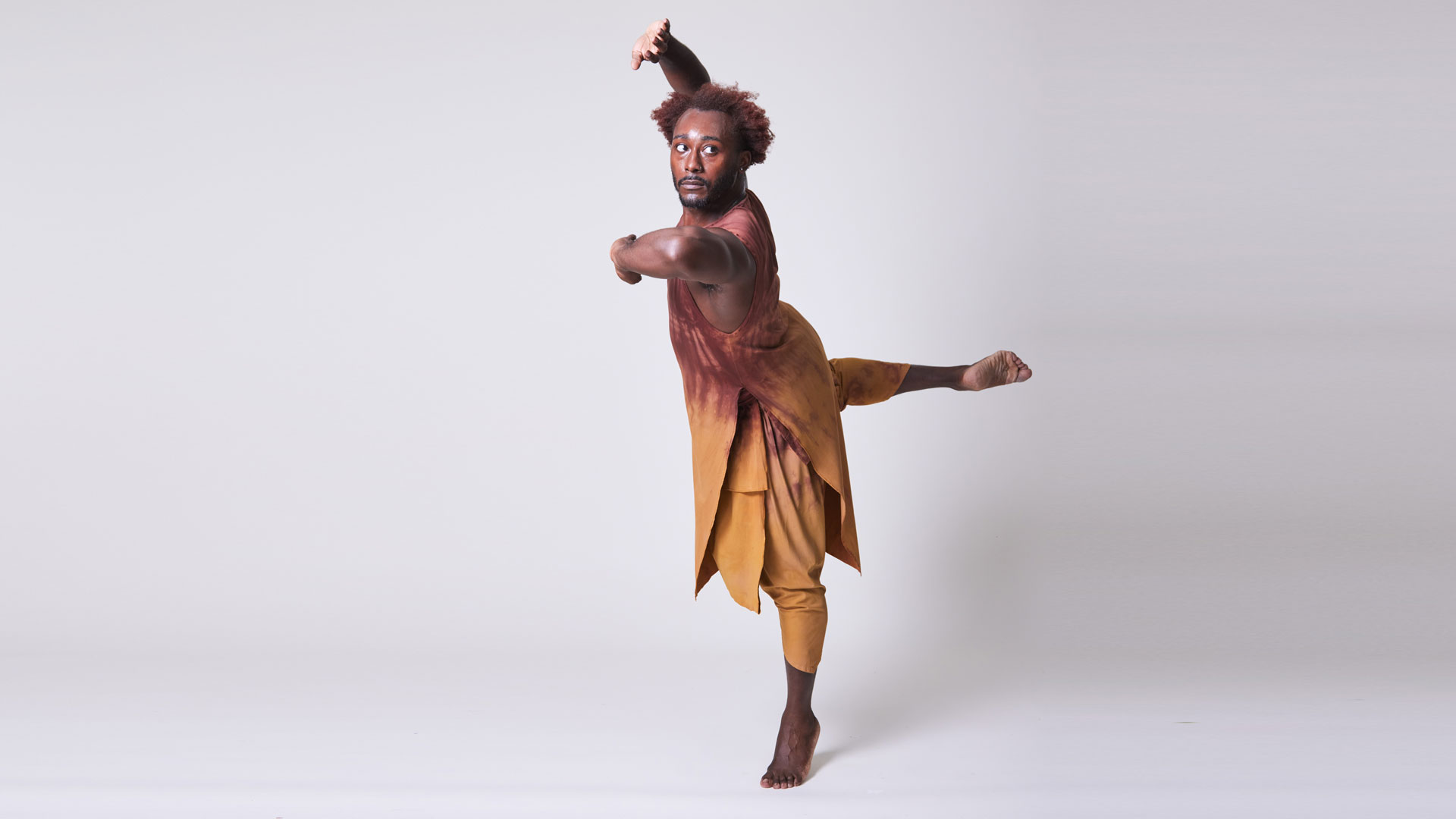 A Black dancer with brown hair and a beard wears a warm yellow and brown tunic and pants. They stand on one foot in releve with the other leg in a back attitude. The dancer holds their front arm to their chest and their other arm over their head.