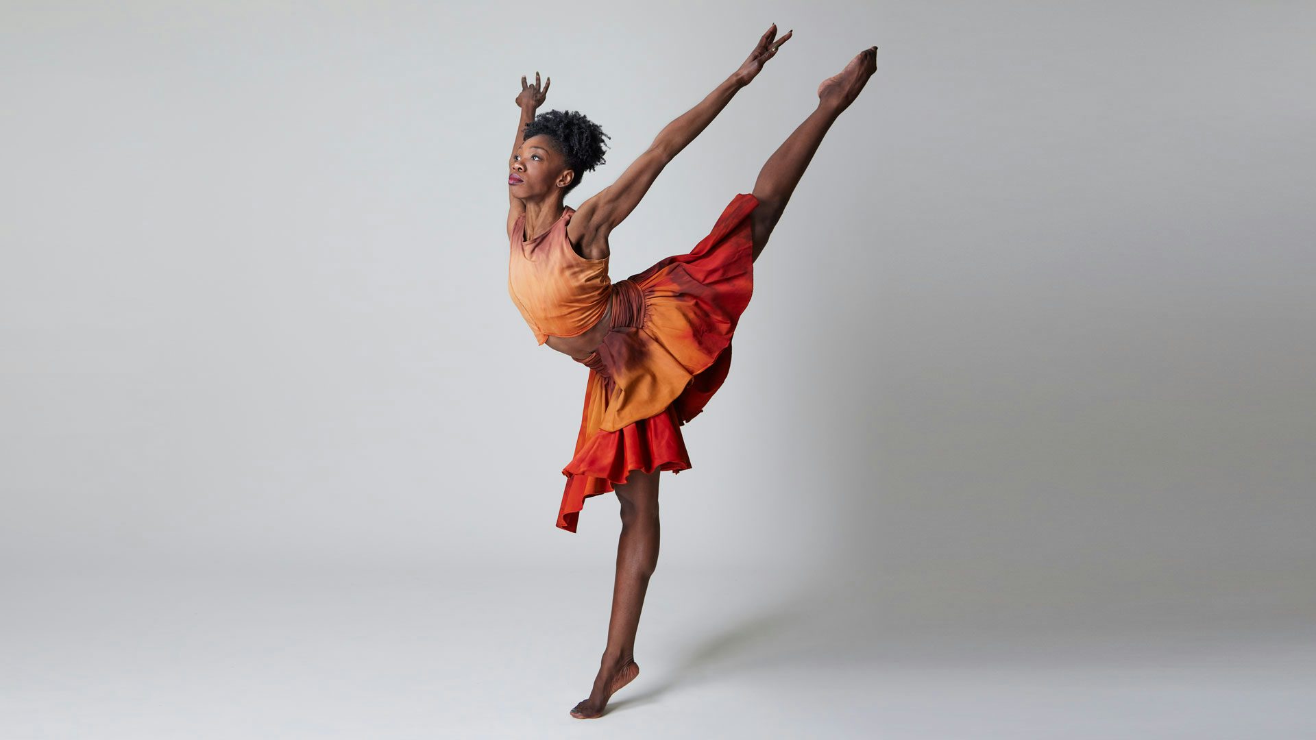 A Black dancer with her dark hair pulled up wears an orange, brown, and red tank top and knee-length layered skirt. She stands on one foot in releve with the other leg held straight behind her with pointed toes. She lifts her arms high behind her in a raised V shape.
