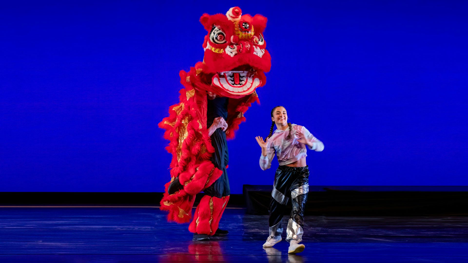 A dancer with brown hair in two plaits wearing a blue and purple tie-dye hoodie and black and silver pants stands on a stage with a blue background. She steps forward on her right foot and smiles, holding her hands at her chest with palms facing out. A tall, human-sized puppet of a red dragon with yellow accents stands on one leg behind and to the left of the dancer.
