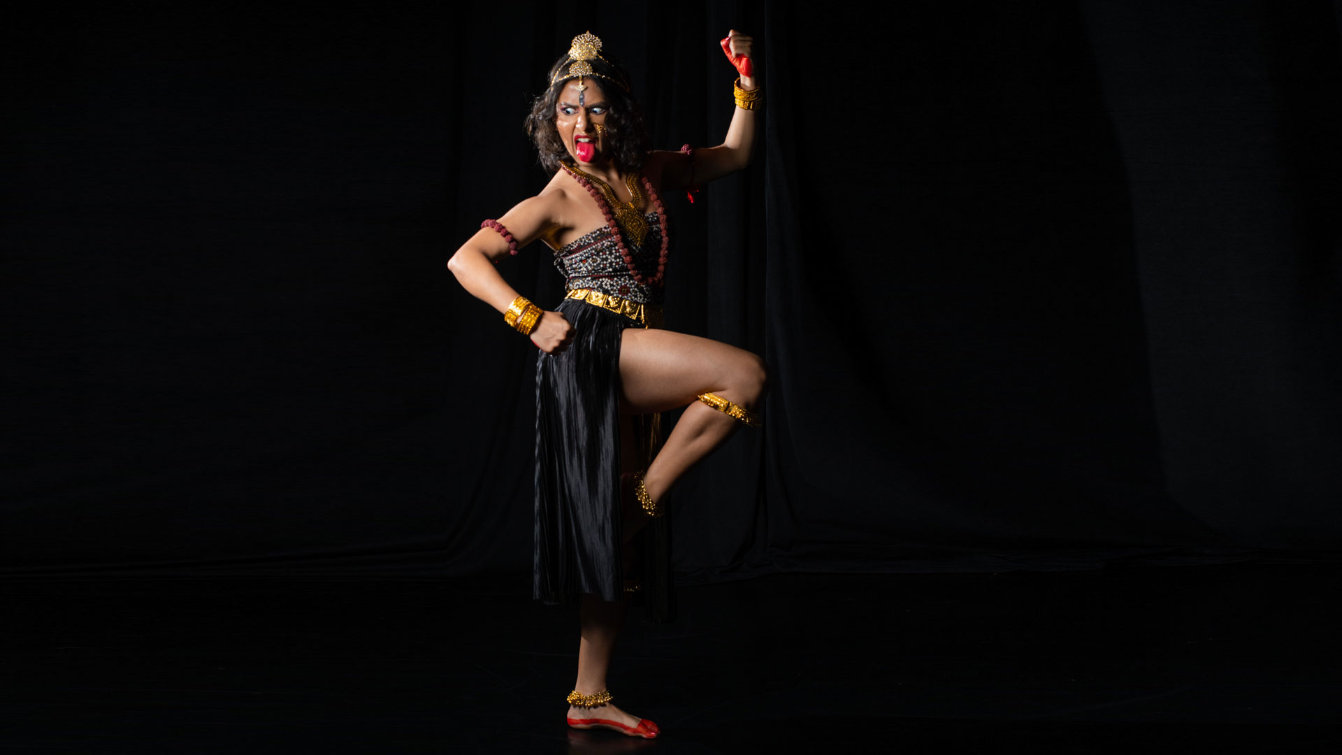 A woman with short brown hair wears a gold headdress, nose ring, necklace, bangles and anklets. Her hands, feet and tongue are painted a bright red. She stands on one foot and lifts a bent leg with foot flexed. The woman holds one bent arm at her hip and one arm bent upward. The woman looks down and left and sticks out her tongue.