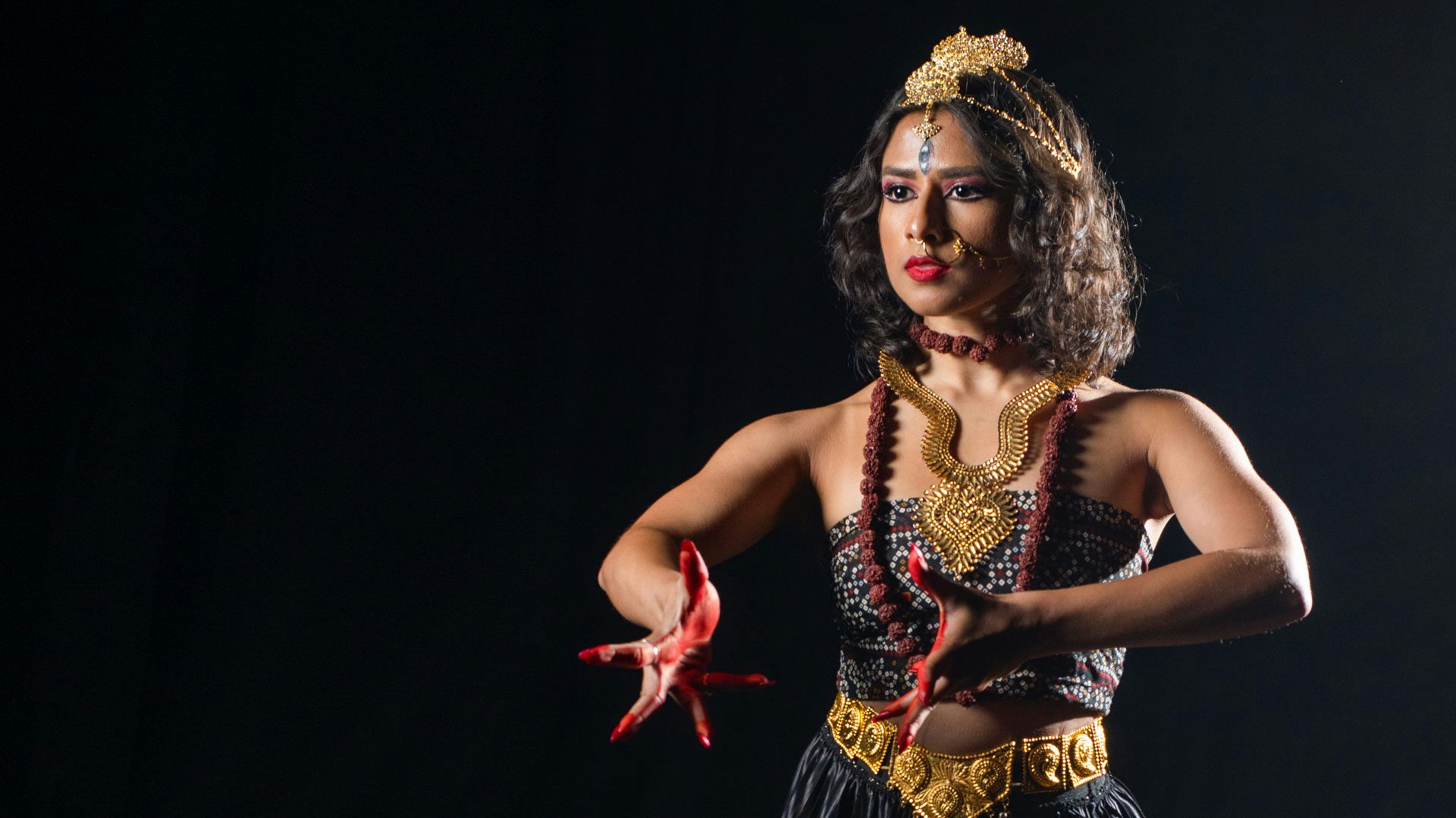 A woman with short brown hair wears a gold headdress, nose ring, belt and necklaces. Her hands and lips are painted bright red. She wears black eyeliner and maroon eyeshadow. There is a sideways eye painted on the center of her brow. The woman bends both arms and holds both hands in front of her.