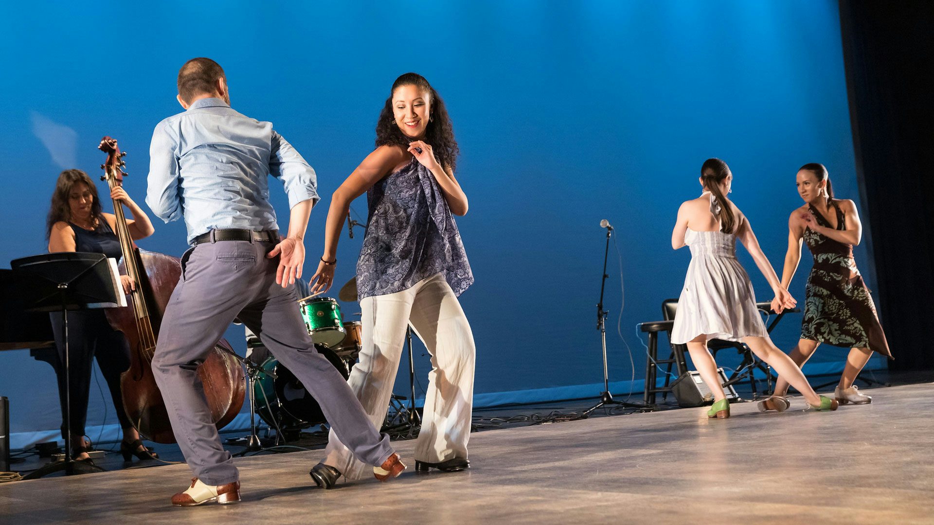 Four dancers wearing tap shoes stand in groups of two on either side of a stage with a sky blue backdrop. The dancers on the left side of each pair face away from the audience. In the background, musicians play a double bass and a green drum set.