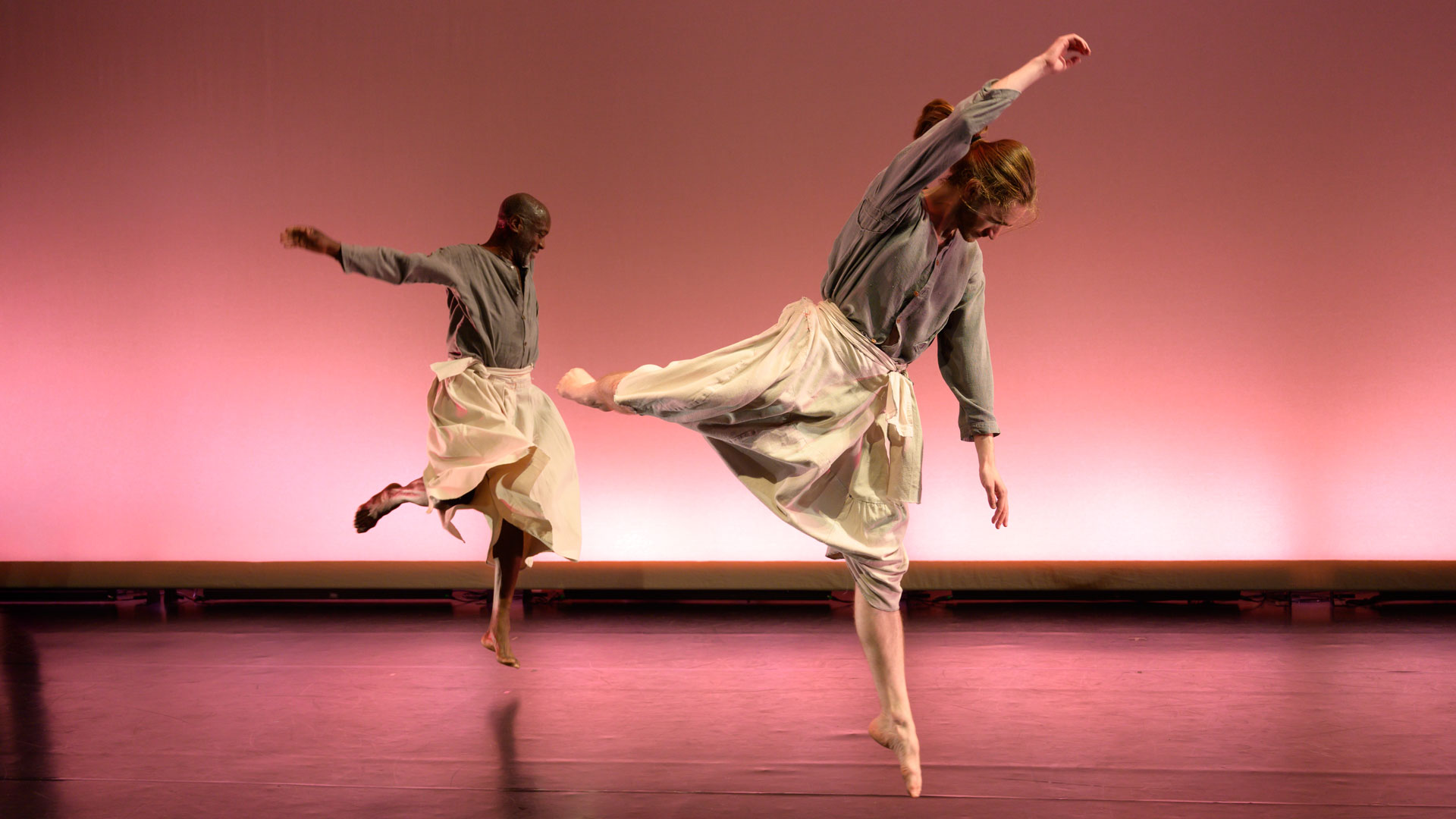 Two dancers wear loose-fitting grey shirts and cream pants. One dancer is slightly closer to the camera than the other. Facing the side of the stage, the dancers leap up on one foot with their opposite arm raised, brushing their other foot behind them with a bent knee.