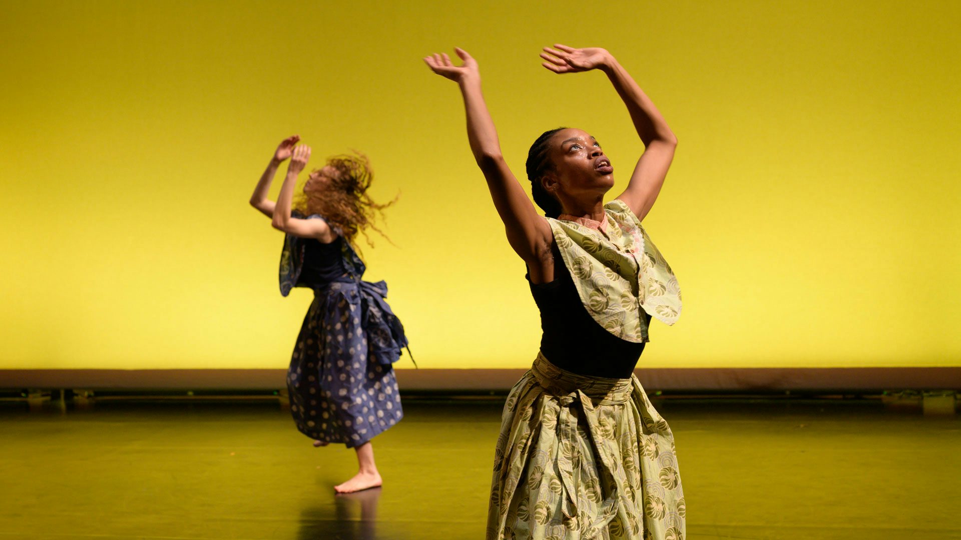 Two dancers raise their arms above their heads and look up towards the ceiling against a bright yellow backdrop on a stage. The dancer in the foreground wears a black tank top under a green patterned loose-fitting skirt and button-up top. The dancer in the background is barefoot and wears a dark tank top under a deep blue loose-fitting skirt and top.