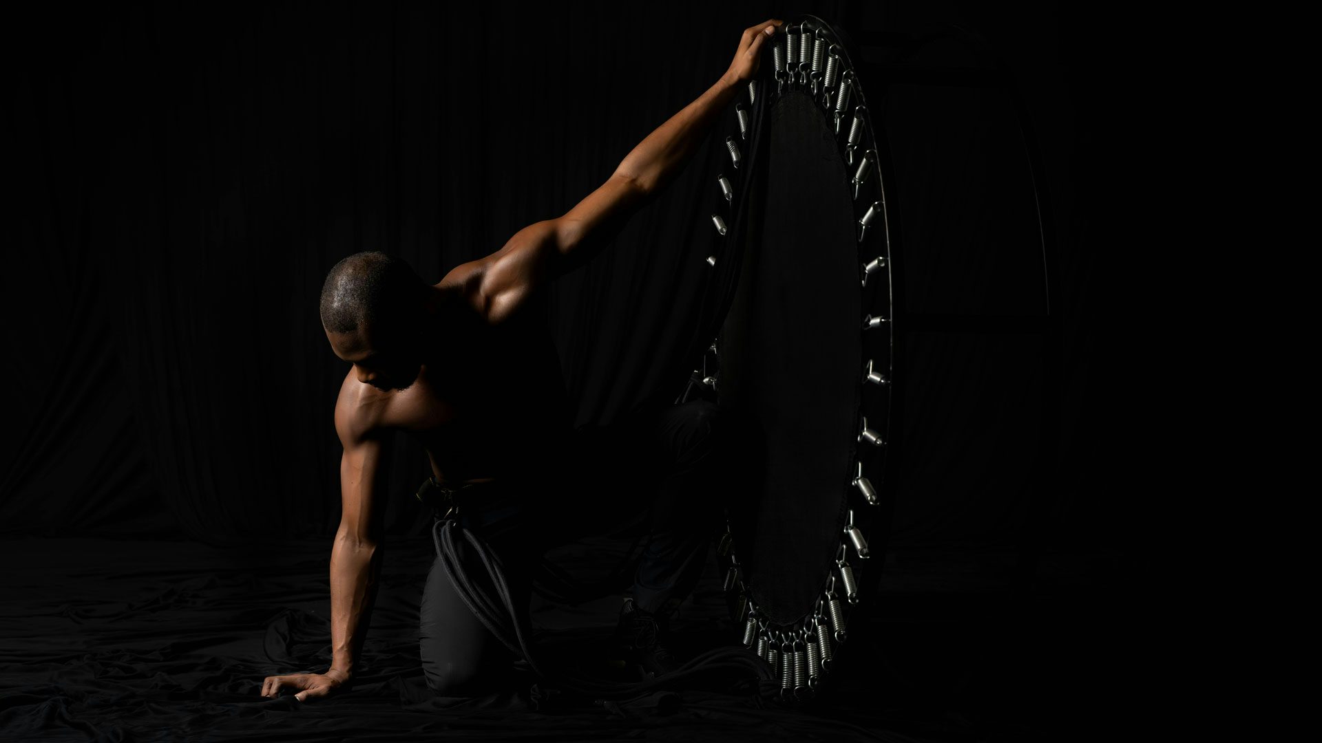 A Black man wearing black pants against a dark background in low light kneels on a floor with both knees and looks down and to the left. He rests one hand on the ground and holds the top of a sideways trampoline with the other hand.