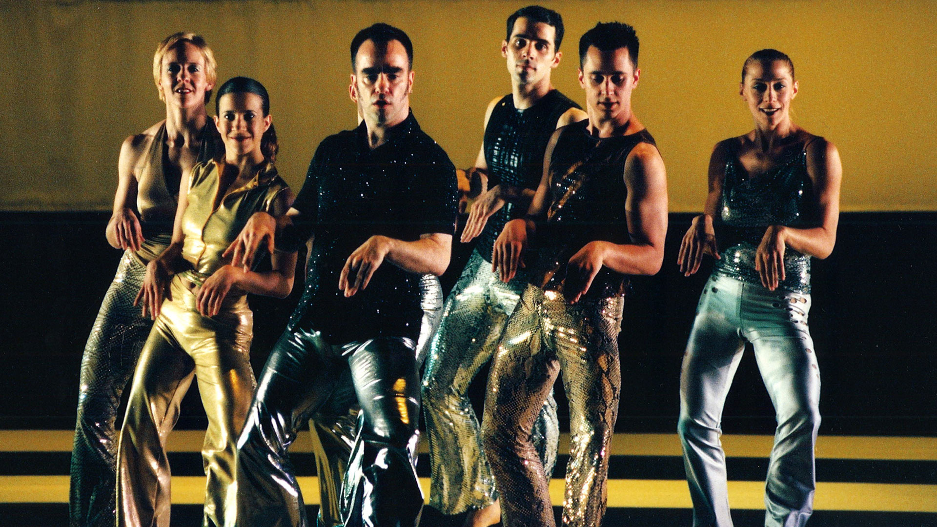 Six dancers in shiny metallic clothing dance in a pyramid formation on a stage against a gold background. The dancers hold their arms at a right angle in front of them, palms facing down, fingers together.