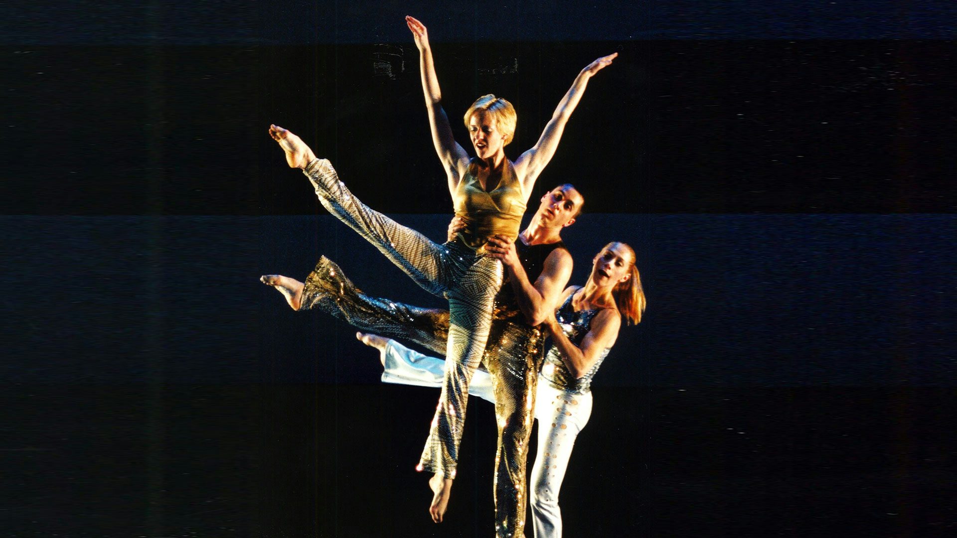 Three dancers in shiny metallic tank tops and flared pants stand in a line in front of each other on a black stage. The dancers kick one leg out straight to the side. The dancer in the back wears a ponytail and holds the waist of the dancer in front of her. The second dancer holds the waist of the dancer in front of him and lifts her into the air. The third dancer in the front holds her arms in a V shape above her head as she is lifted into the air.