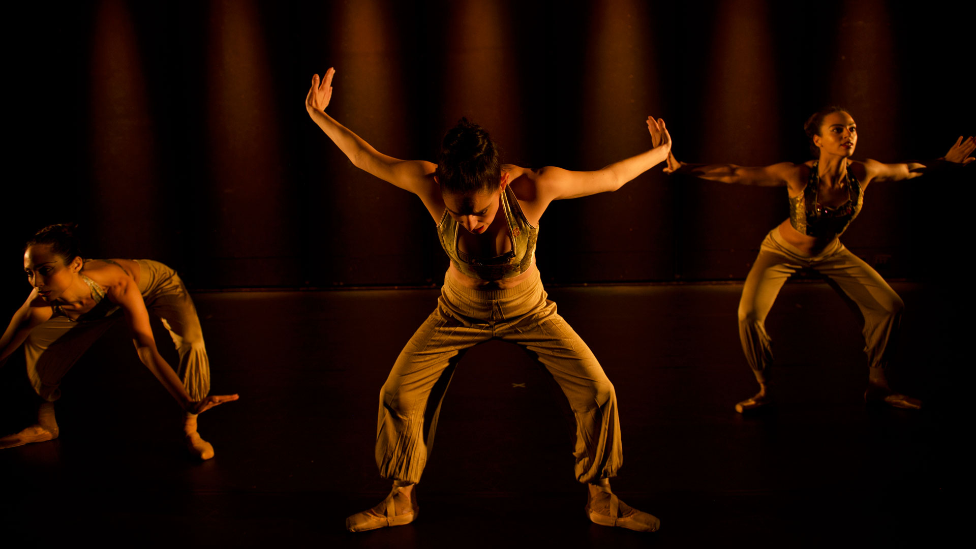 Three women wearing pointe shoes, gold tank tops and pants stand on a dimly lit stage with their feet apart and knees bent. Their arms are raised straight out to their sides, palms flexed away from their bodies.