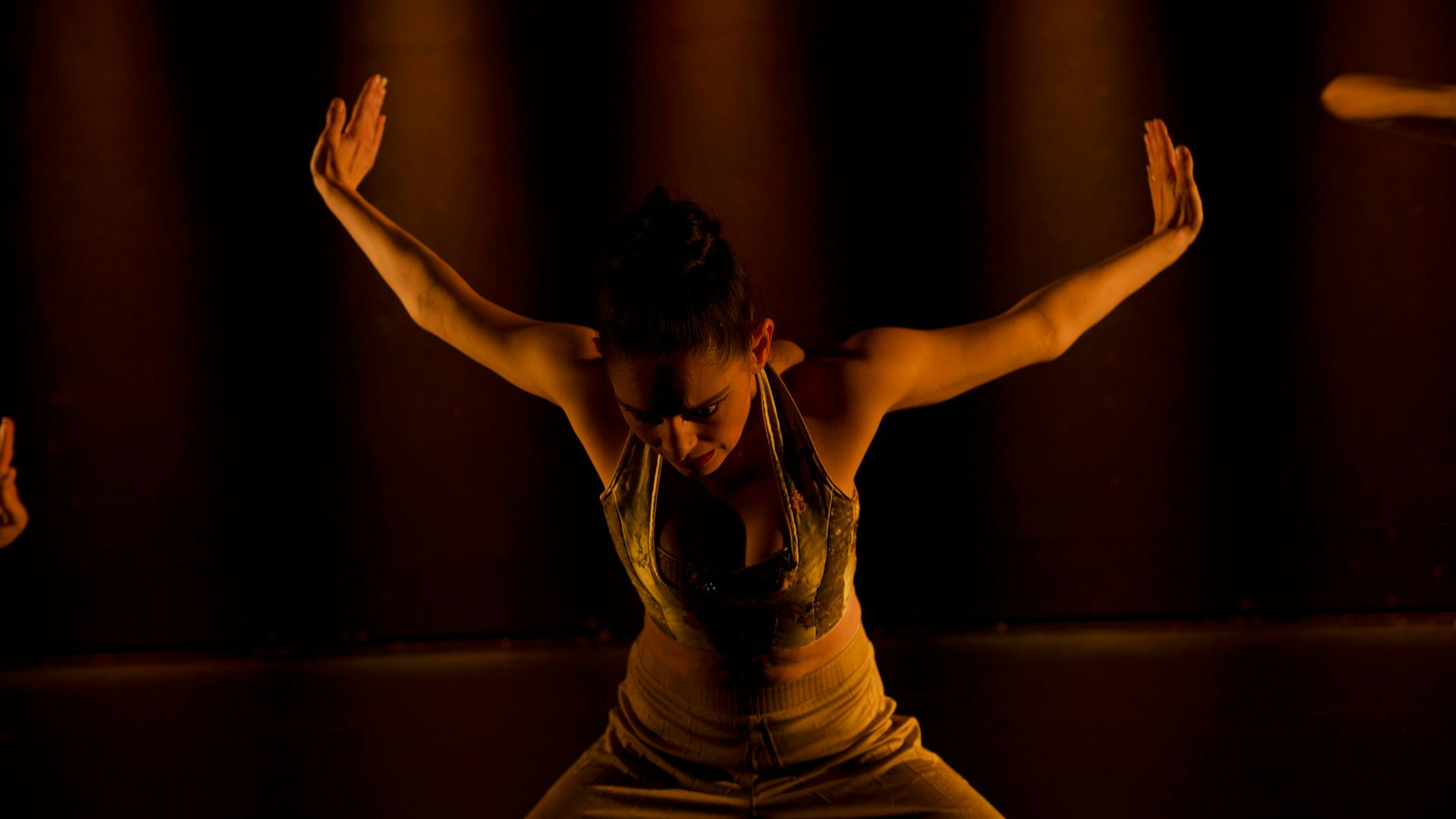 A woman with dark hair in a bun wears and a velvet tank top. She stands on a dimly lit stage with her knees bent and raises her arms straight out behind her, palms flexed away from her body.