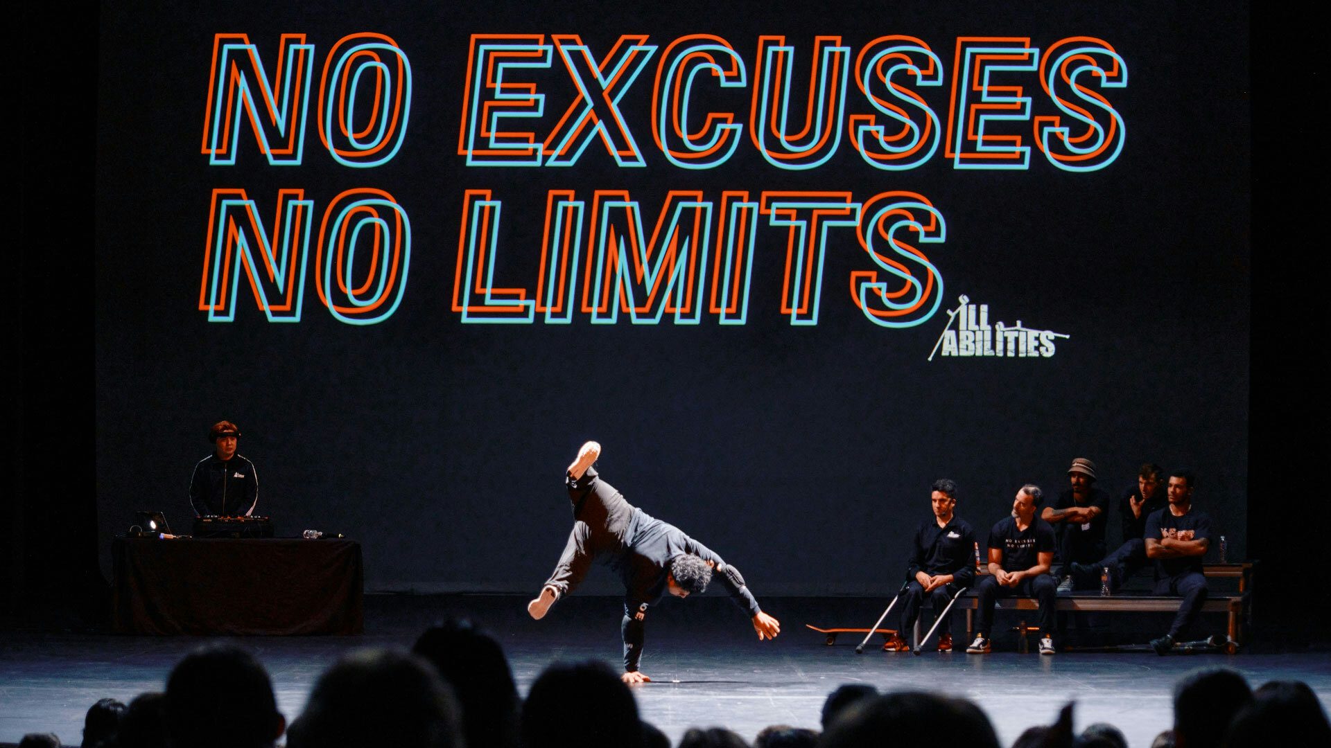 A backdrop reads “No Excuses, No Limits.” On stage, a dancer balances on one hand with his legs spread out in the air. Behind him, a DJ wearing headphones stands at a turntable.