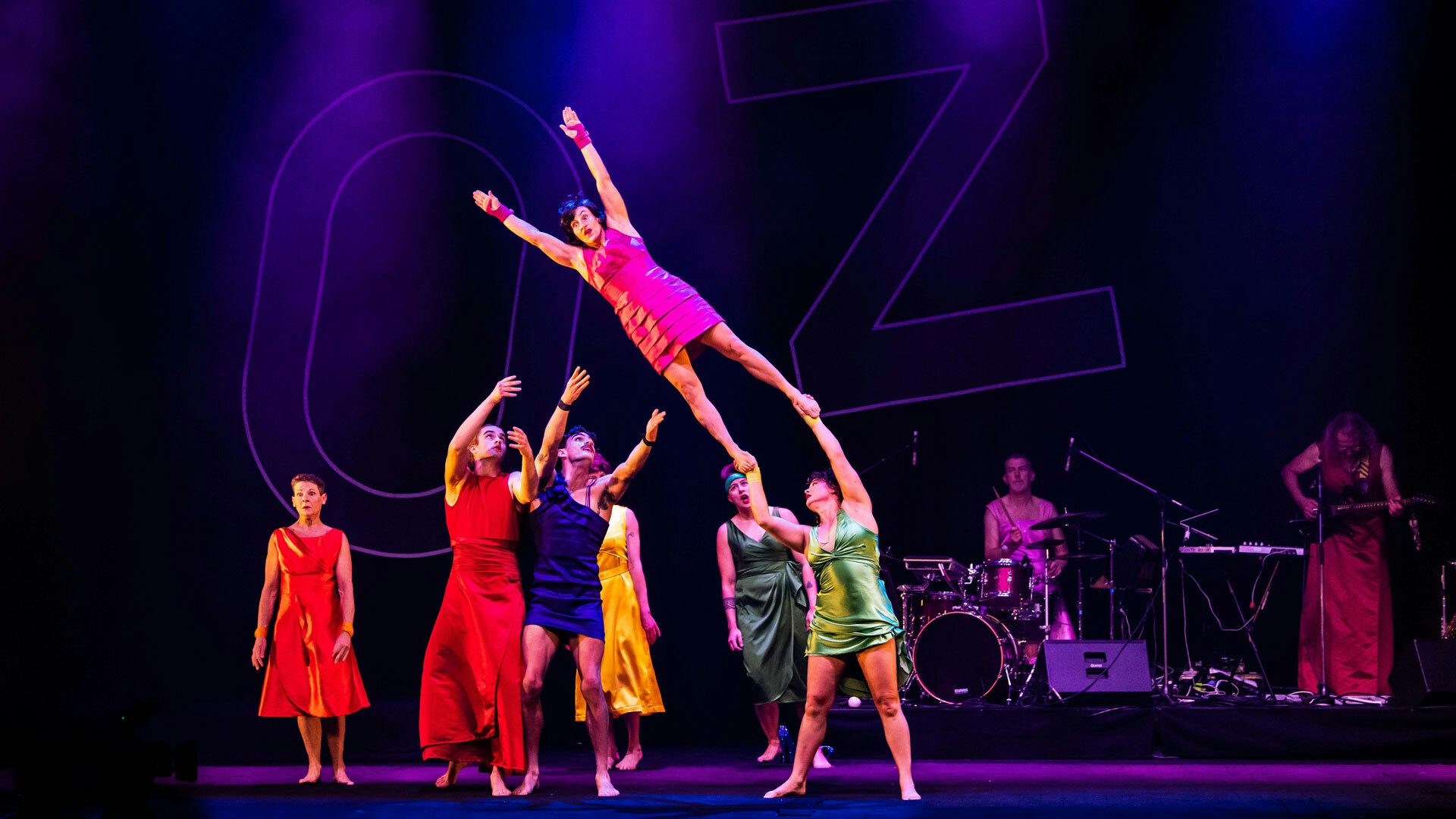 One performer in a bright pink dress flies above the others as a performer in a lime green dress holds their feet and two other performers catch them.